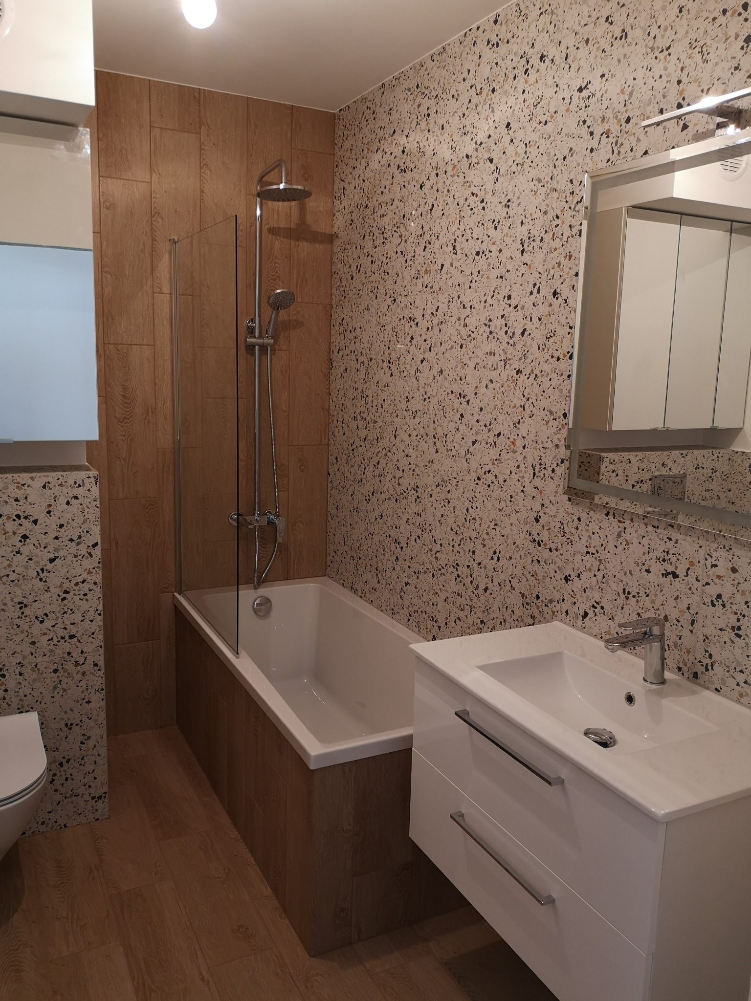 Nowoczesna łazienka po remoncie w Olesno: wanna z parawanem, umywalka z szafką i mozaikowa ściana. Drewnopodobne płytki na podłodze i obudowie wanny.