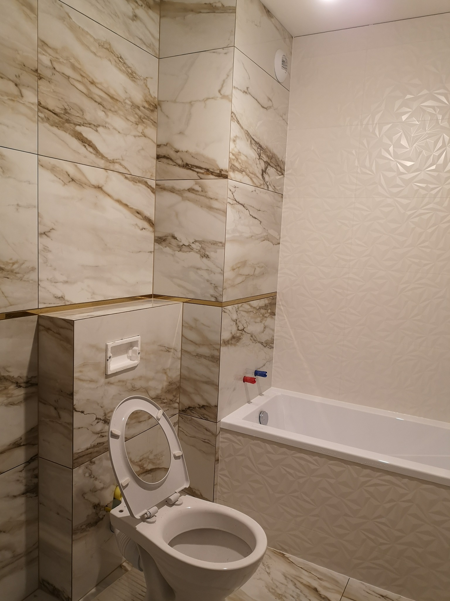 Elegancka łazienka po remoncie: marmurowe płytki z złotymi akcentami, geometryczne wzory na ścianie przy wannie, otwarta klapa toalety. Nowoczesny design.