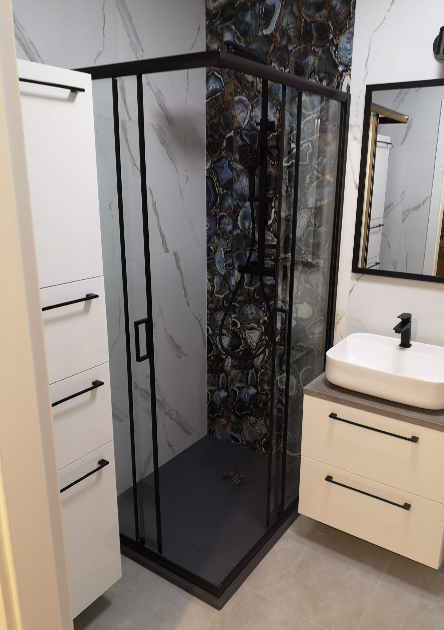 Nowoczesna łazienka z kabiną prysznicową typu walk-in z czarnymi profilami, umywalką nablatową i szafkami w kolorze złamanej bieli. Na ścianie płytki imitujące marmur i agat.