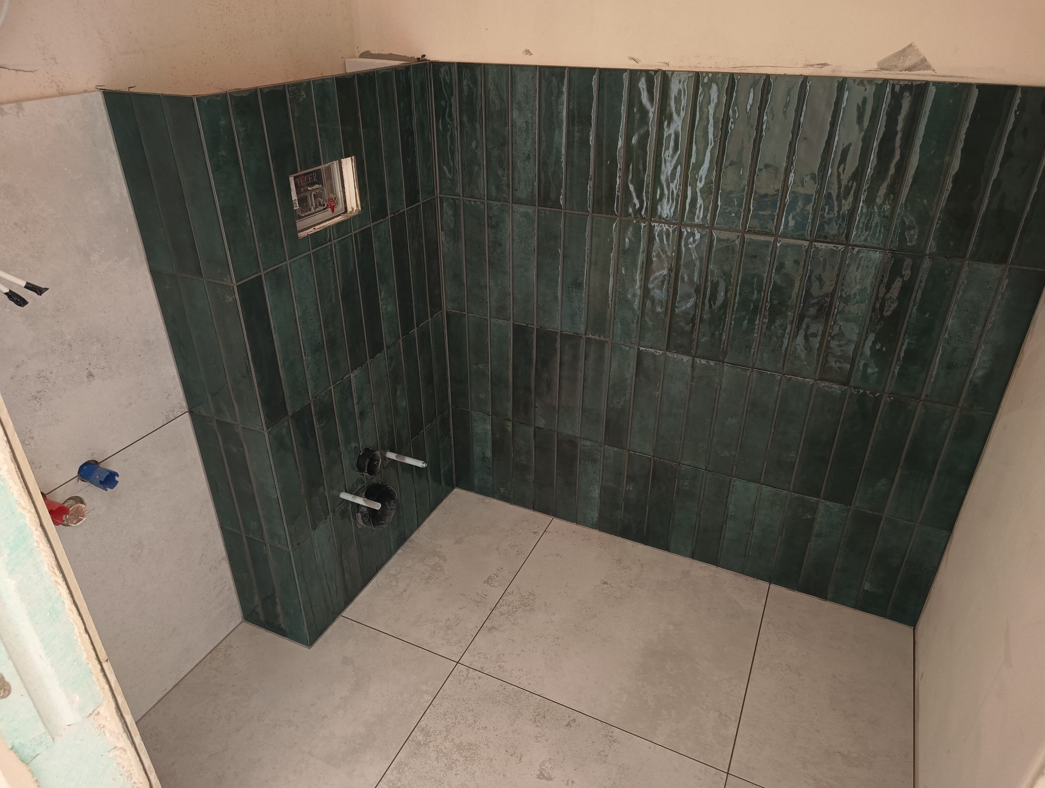 Łazienka po remoncie: ściany wyłożone pionowymi, ciemnozielonymi płytkami o błyszczącej fakturze. Na podłodze jasne, kwadratowe płytki. Widoczne przyłącza wodne.