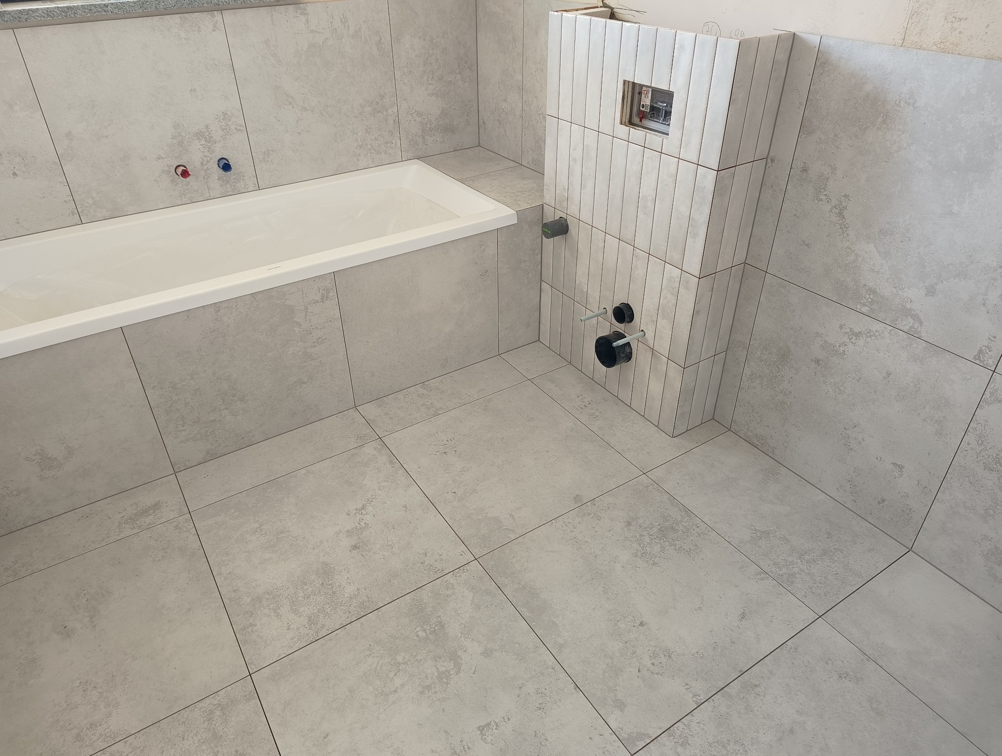 Wykończona łazienka z wanną obłożoną szarymi płytkami. Widoczne pionowe płytki na zabudowie stelaża WC i podłączenia hydrauliczne. Minimalistyczny design.