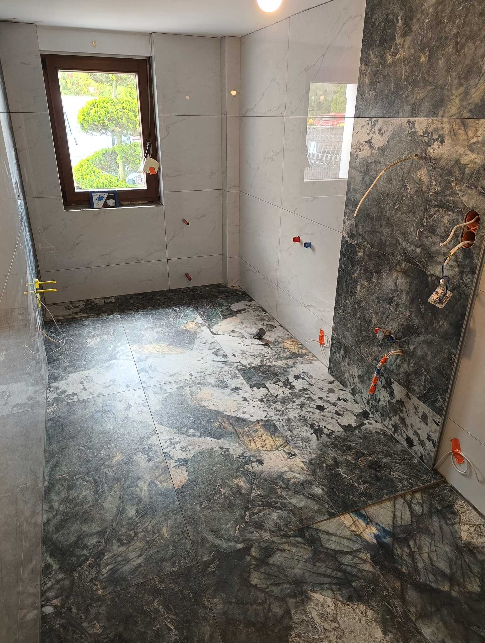 Łazienka w trakcie wykańczania: położone płytki podłogowe o wzorze marmuru, białe płytki ścienne, widoczne instalacje elektryczne i hydrauliczne, okno z widokiem na zieleń.