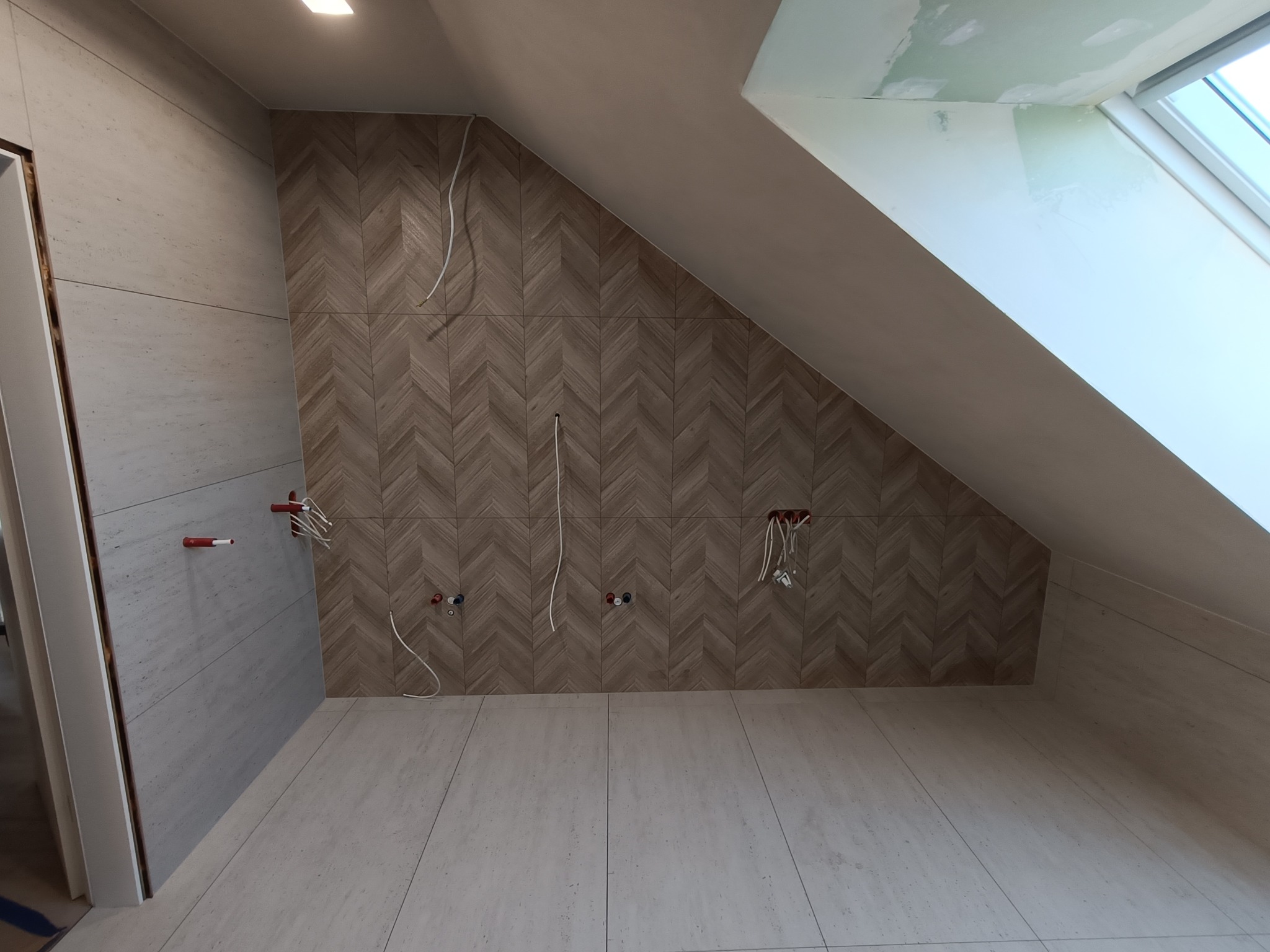 Łazienka na poddaszu z wykończoną ścianą w jodełkę z płytek, widoczne instalacje hydrauliczne i elektryczne, okno dachowe. Stonowana kolorystyka.