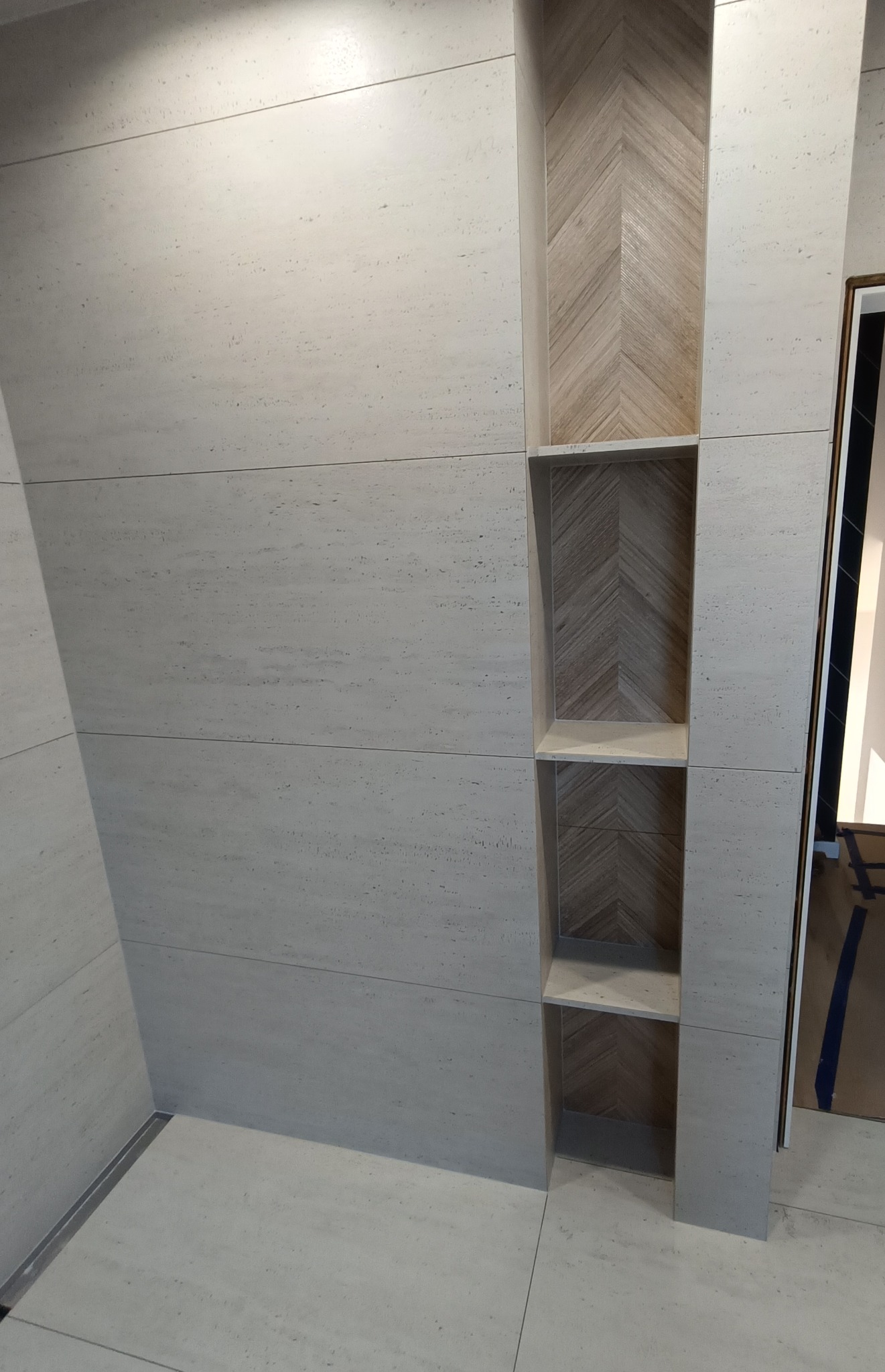 Wykończona ściana w łazience z wnęką wyłożoną drewnopodobną płytką w jodełkę. Widoczny odpływ liniowy w podłodze i fragment drzwi.