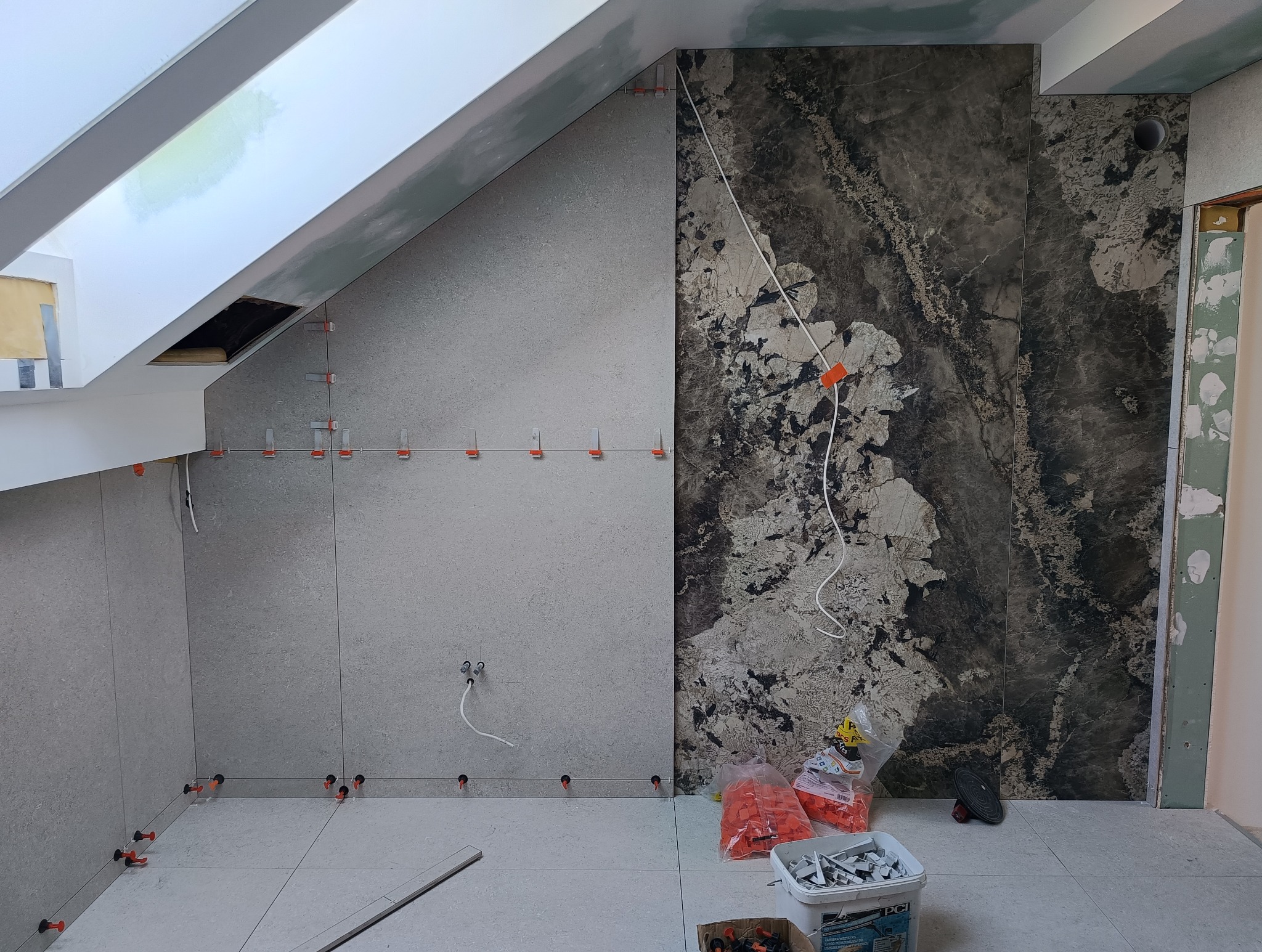 Łazienka w trakcie wykańczania: szare płytki na ścianach i podłodze, ciemna ściana z wzorem imitującym kamień, system poziomowania płytek. Skos dachu.