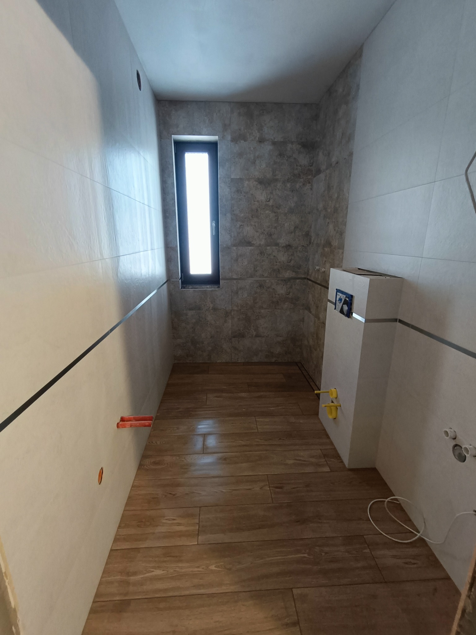 Wykończona łazienka z drewnopodobną podłogą, jasnymi płytkami na ścianach i oknem. Widoczna zabudowa WC i instalacje hydrauliczne.