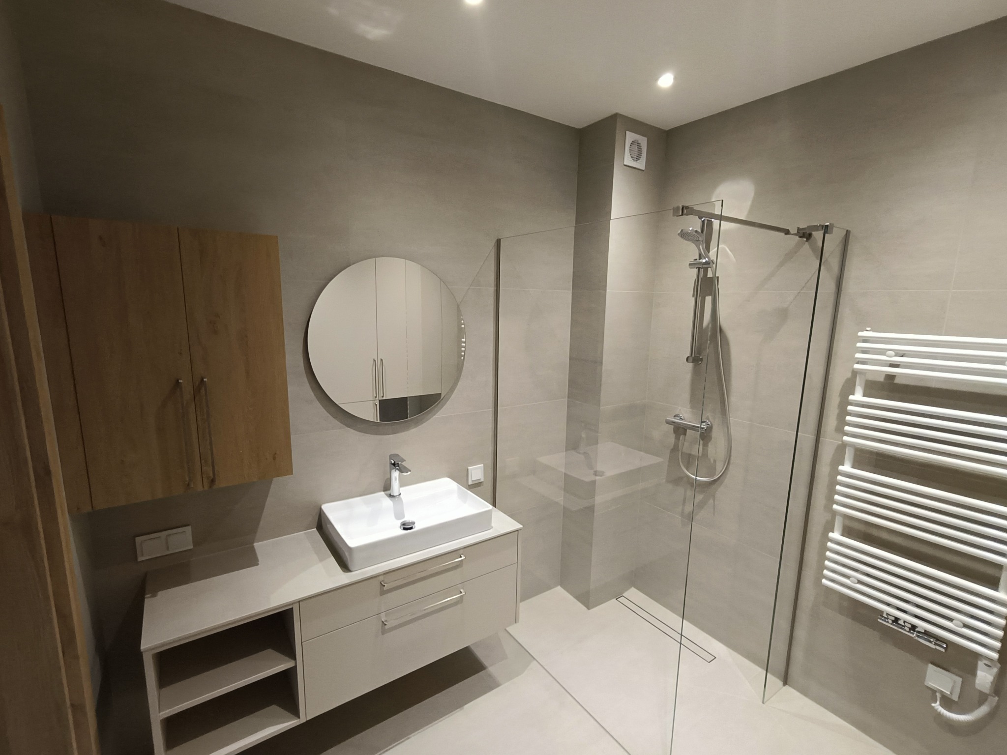 Nowoczesna łazienka w odcieniach szarości z drewnianą szafką. Kabina prysznicowa ze szklaną ścianką, okrągłe lustro i biała umywalka z szafką podwieszaną.