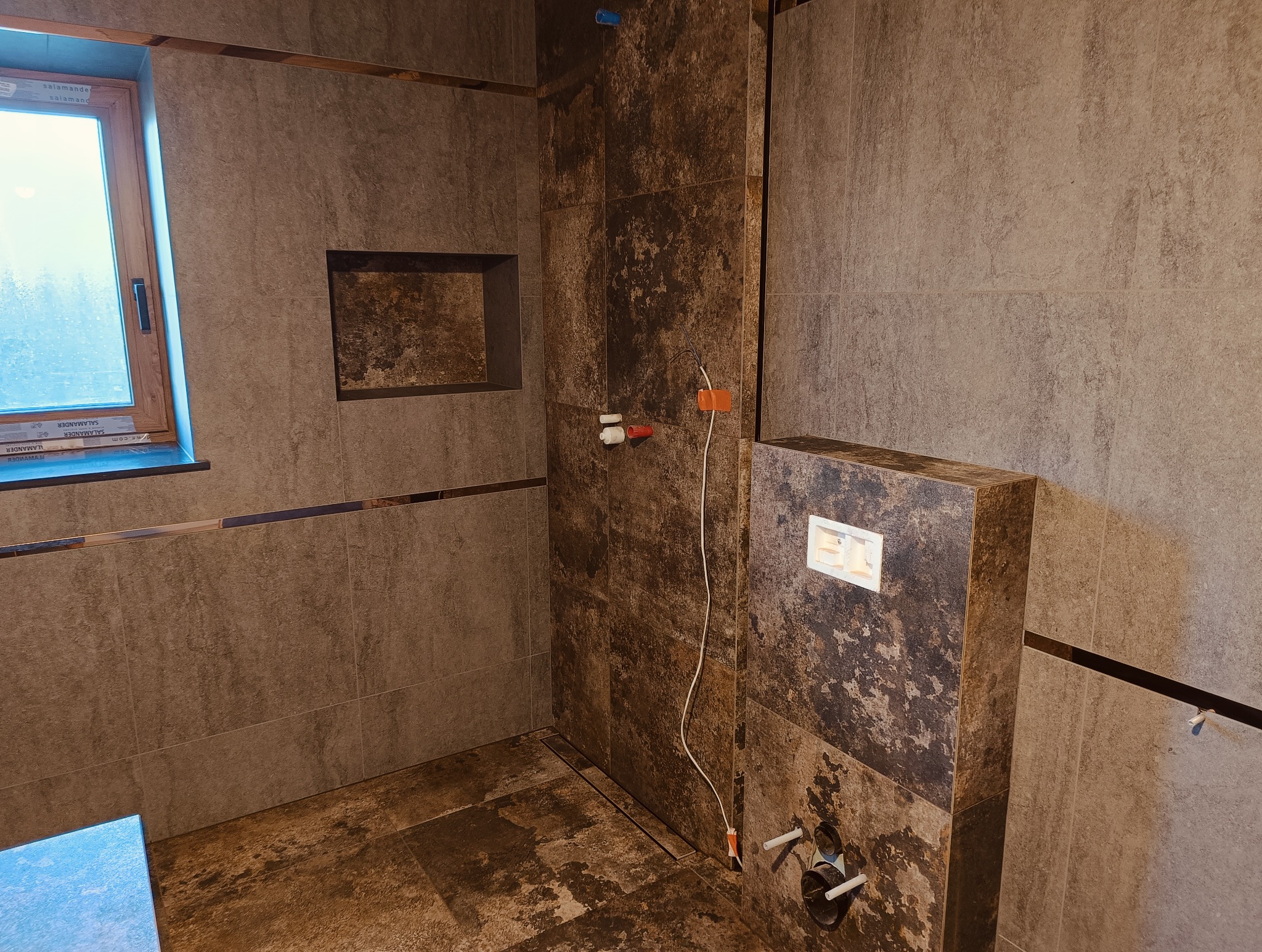 Stylowa łazienka w trakcie wykańczania: brązowe płytki imitujące kamień, podtynkowa spłuczka WC, okno z widokiem na zewnątrz. Widoczne instalacje wodne i elektryczne.