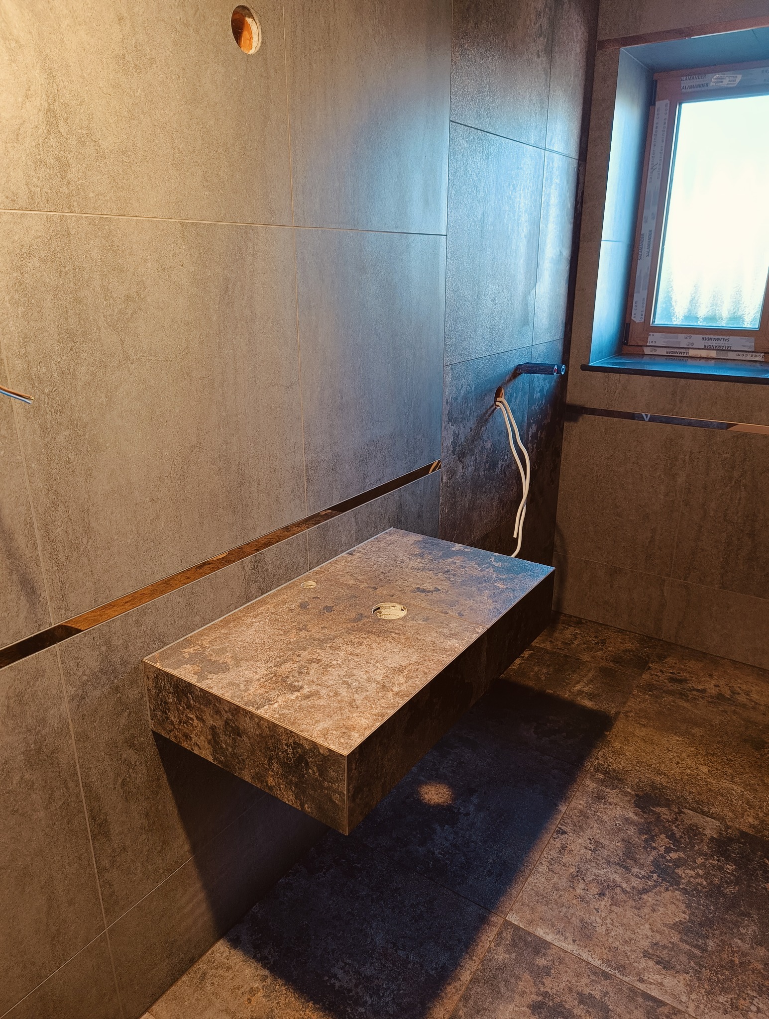 Wykończona łazienka z płytkami w odcieniach brązu i beżu. Umywalka z płytek zawieszona na ścianie, widoczne wystające przewody. Okno z drewnianą ramą.