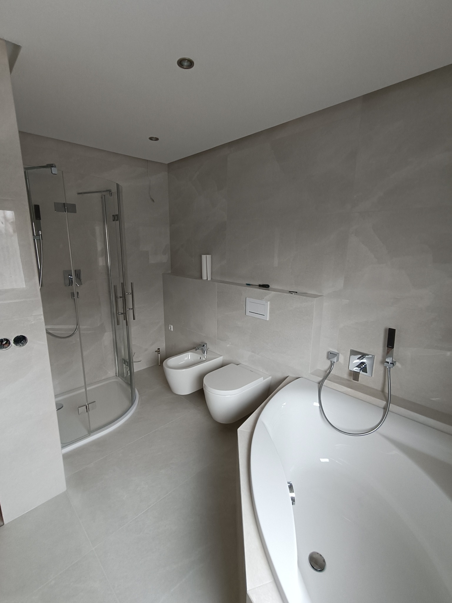 Jasna łazienka po remoncie z kabiną prysznicową, bidetem, toaletą i wanną. Na ścianach i podłodze położone są płytki w odcieniach szarości.