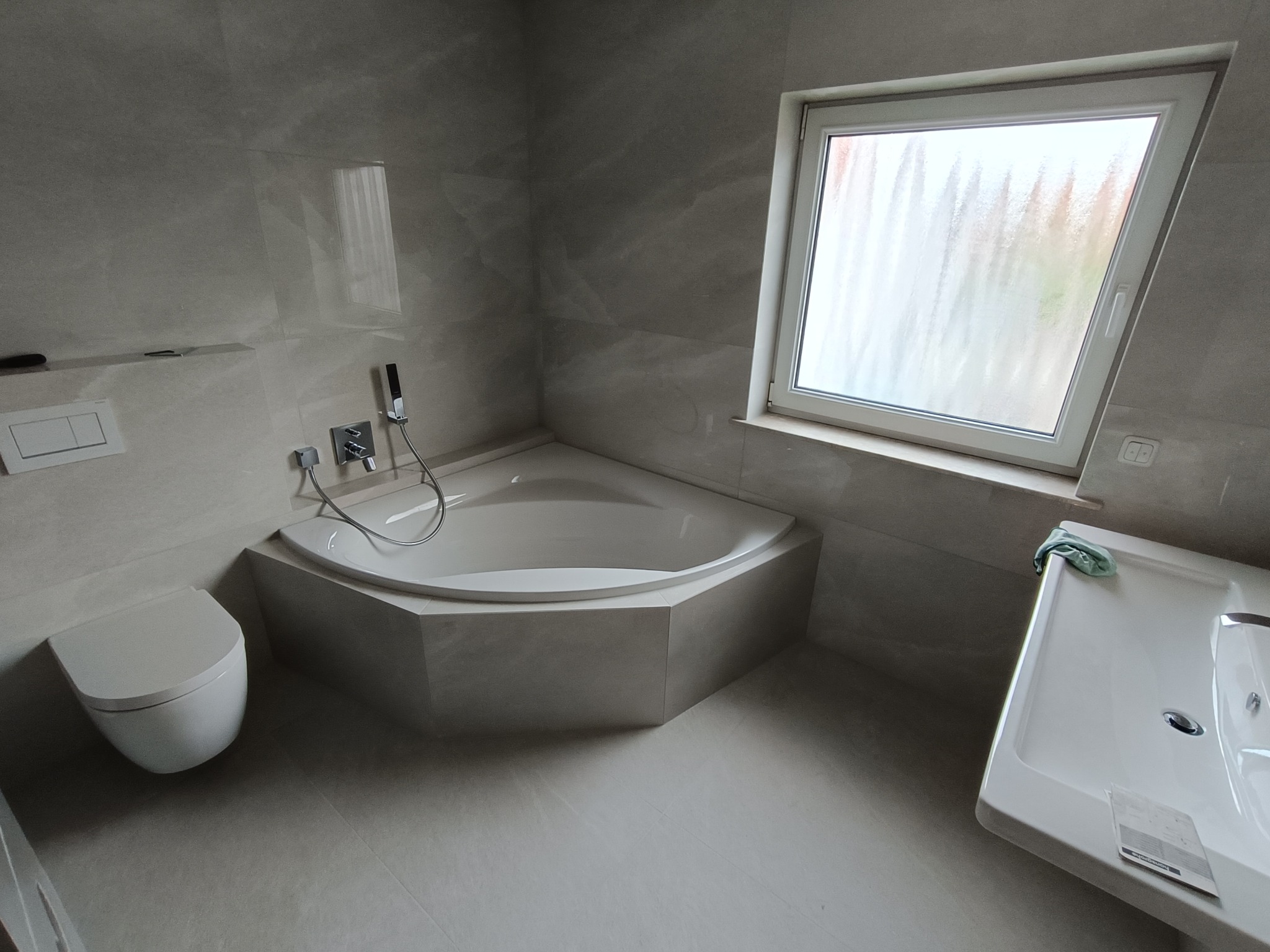 Wykończona łazienka w jasnych odcieniach szarości z narożną wanną, podwieszaną toaletą i umywalką. Okno z rozmytą szybą. Minimalistyczny design.