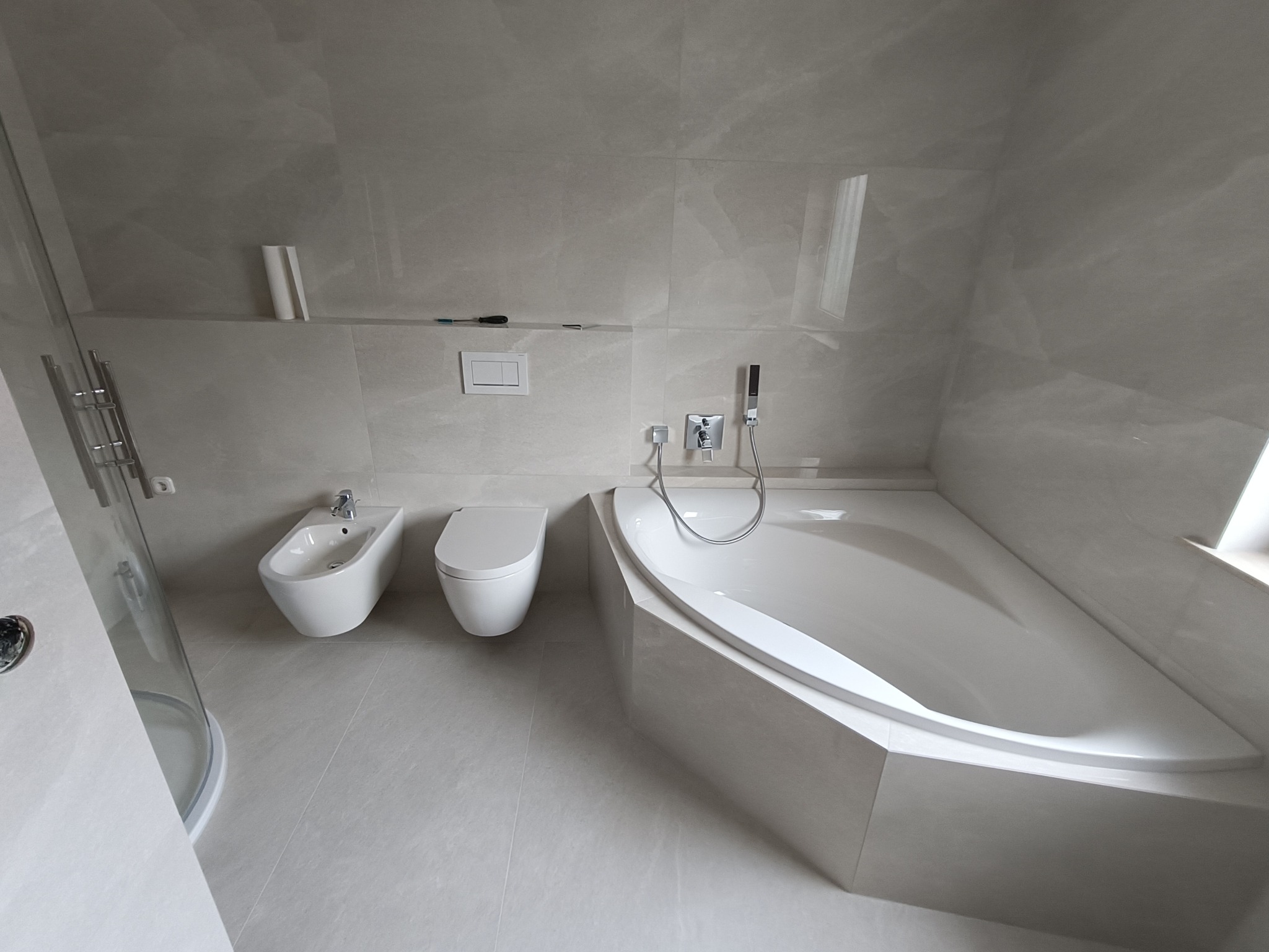 Elegancka, jasna łazienka z wanną, bidetem i toaletą. Płytki imitujące marmur, minimalistyczna armatura. Widoczny fragment kabiny prysznicowej.