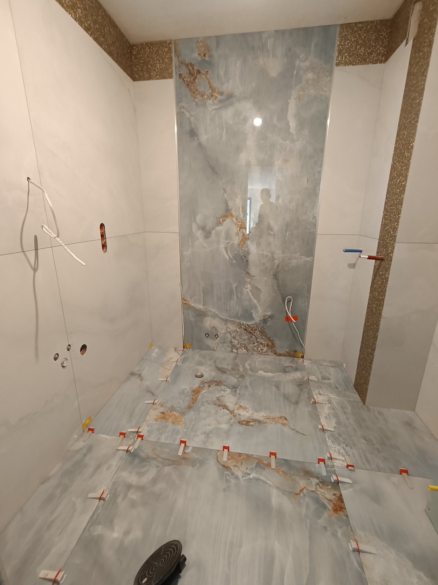 Łazienka w trakcie wykańczania: niebiesko-białe płytki imitujące marmur na podłodze i ścianach, krzyżyki dystansowe, widoczne instalacje. Dekoracyjne mozaiki na górze ścian.