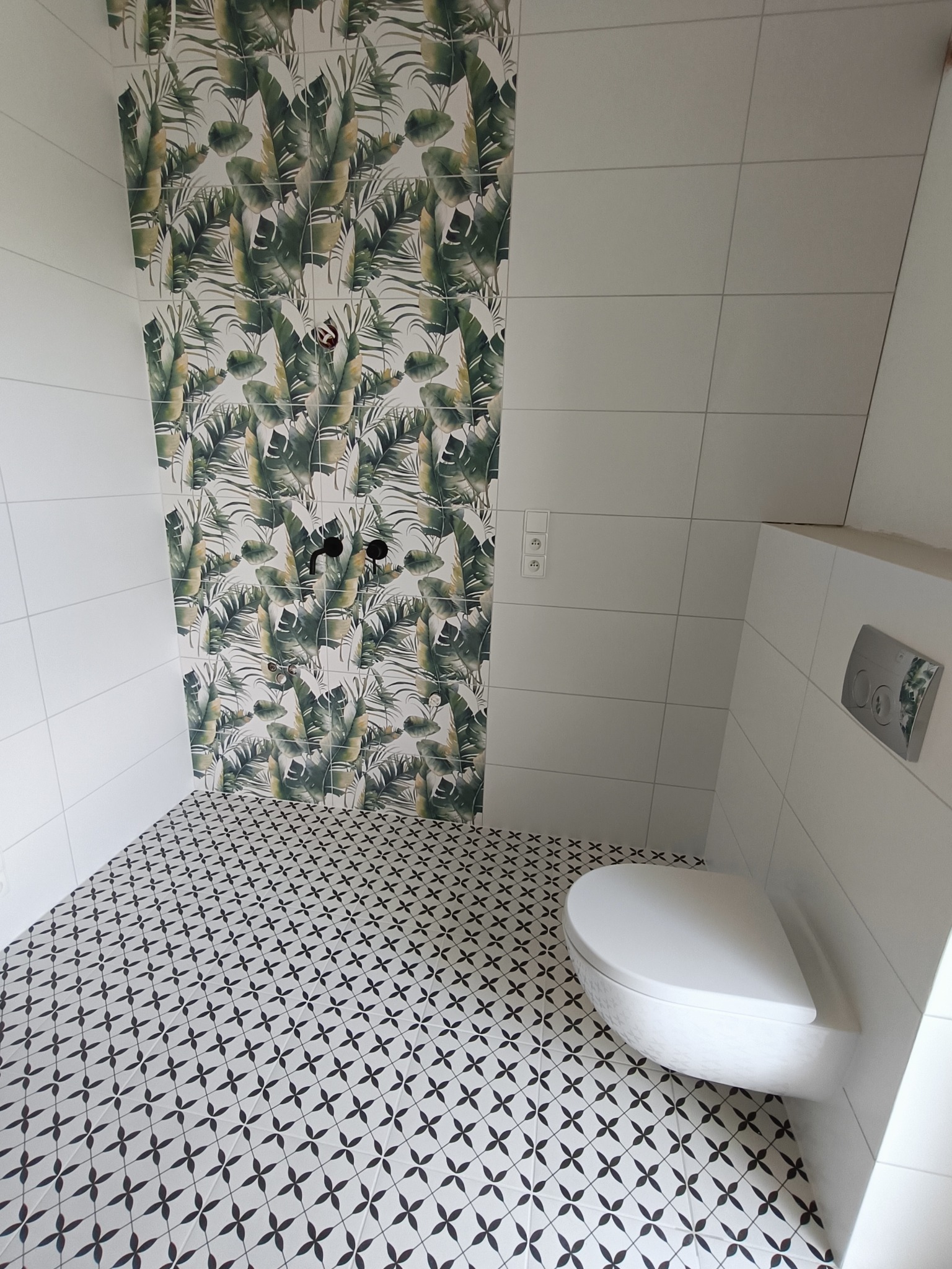 Wykończona łazienka z wzorzystą podłogą, ścianą w tropikalne liście i podwieszaną toaletą. Nowoczesny design z czarną armaturą i białymi płytkami.