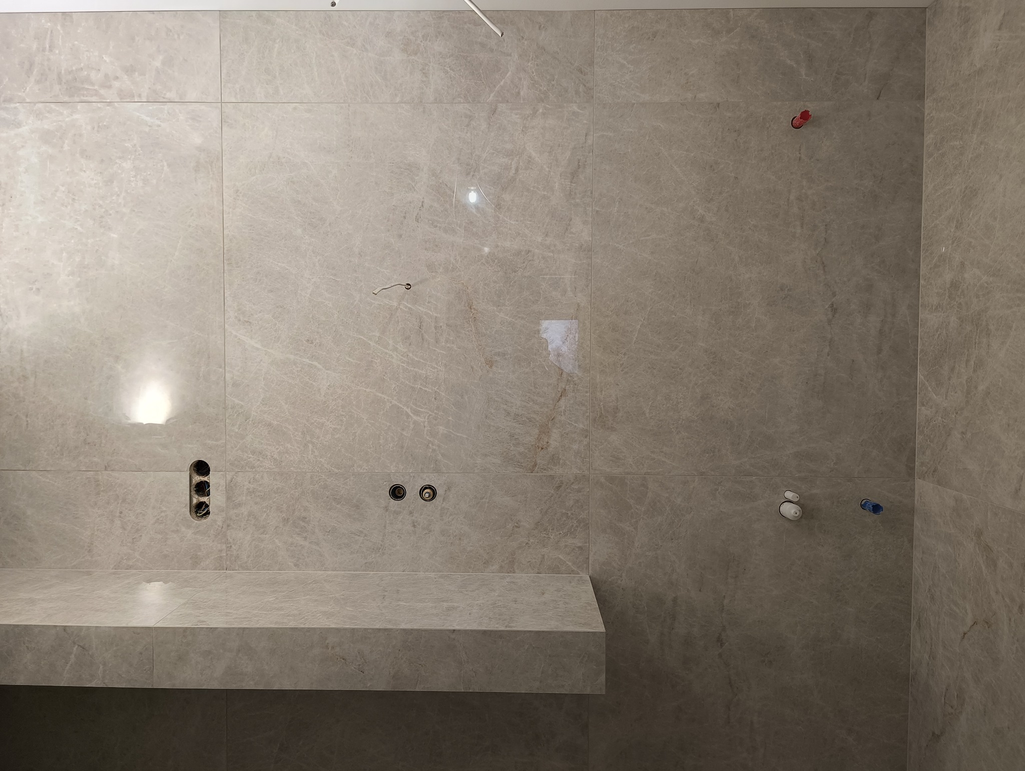 Wykończona ściana łazienki z marmurowymi płytkami w odcieniach szarości, z zamontowanym blatem i przygotowanymi punktami instalacji wodnej i elektrycznej.