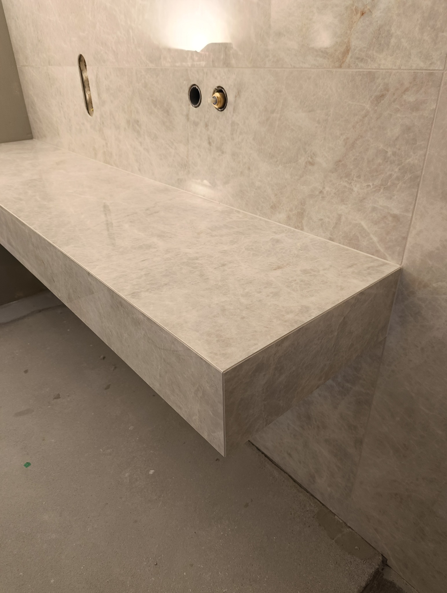 Wykończona, beżowa półka z płytek imitujących marmur w łazience. Widoczne otwory na baterię i odpływ. Minimalistyczny design, surowy beton na podłodze.