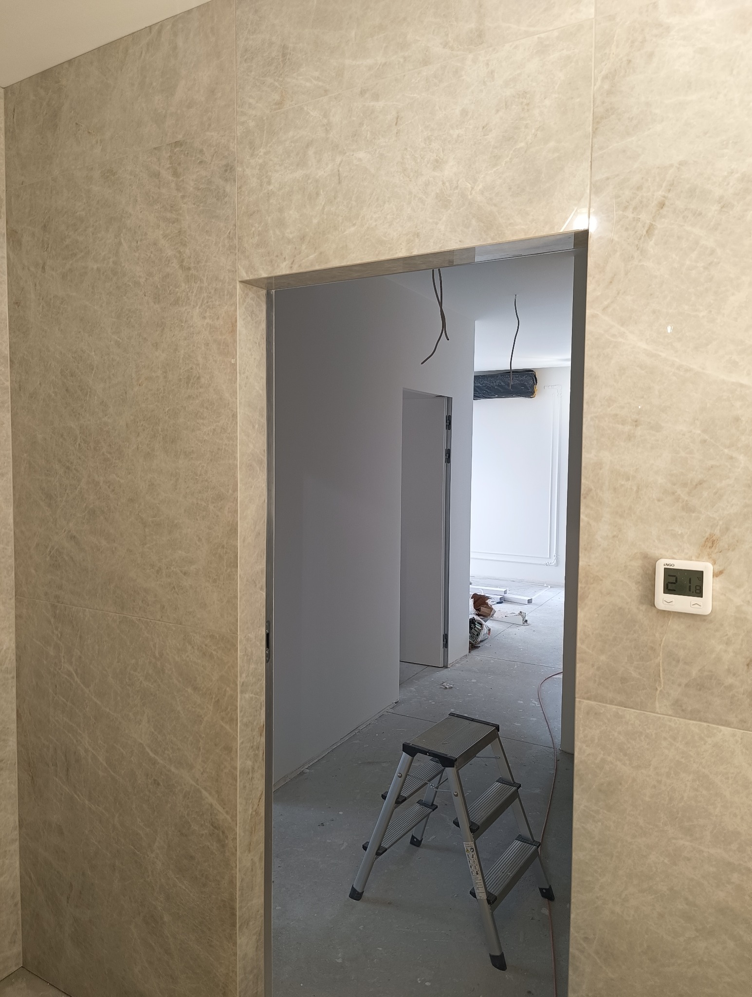 Wykończona ściana z płytek imitujących marmur, widok na wnętrze łazienki w trakcie remontu, drabinka i termostat na ścianie. Neutralna kolorystyka.