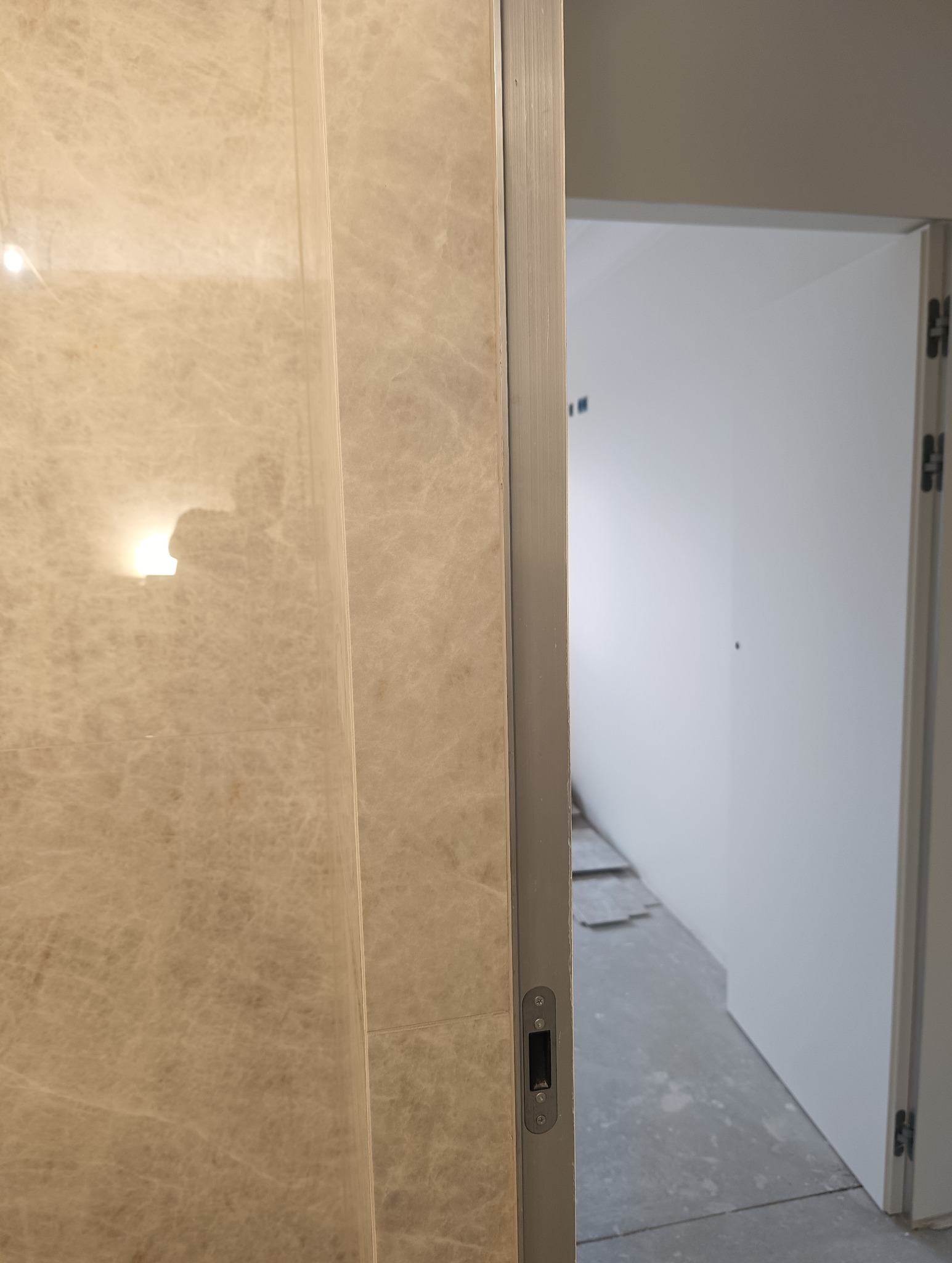 Beżowa, marmurkowa okładzina ściany z widocznym zamkiem drzwiowym i fragmentem wnętrza w tle. Ujęcie z bliska, prezentujące detale wykończenia.