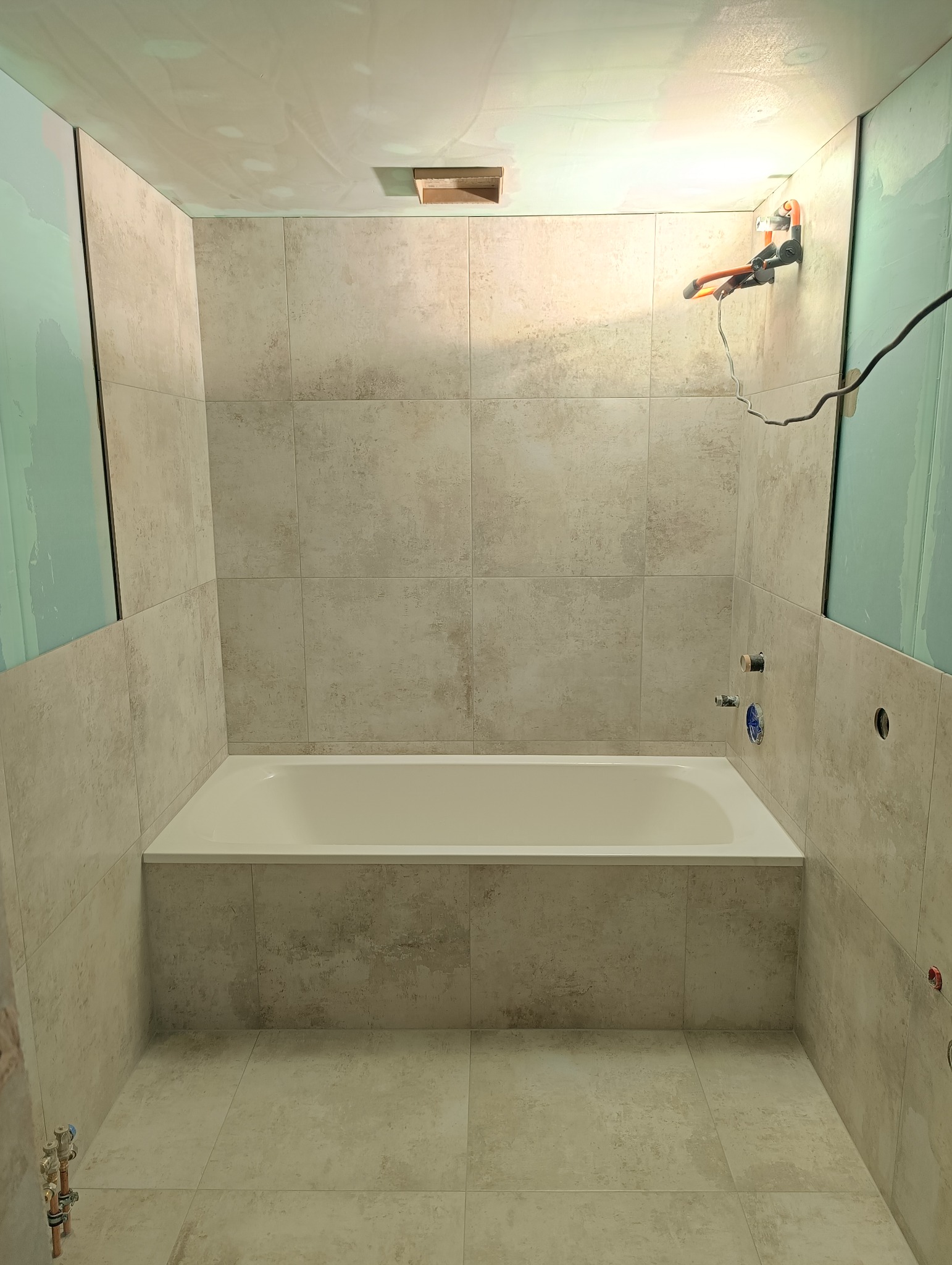 Wykończona łazienka z wanną w zabudowie, ściany i podłoga wyłożone beżowymi płytkami. Widoczne punkty instalacji wodnej i narzędzie do montażu na ścianie.