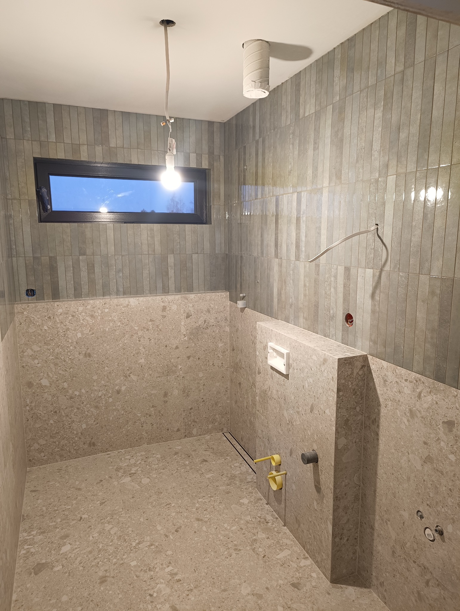 Wnętrze łazienki w trakcie wykańczania: szare, pionowo ułożone płytki na ścianach, beżowa mozaika na podłodze i zabudowie. Widoczne instalacje, okno i wisząca żarówka.