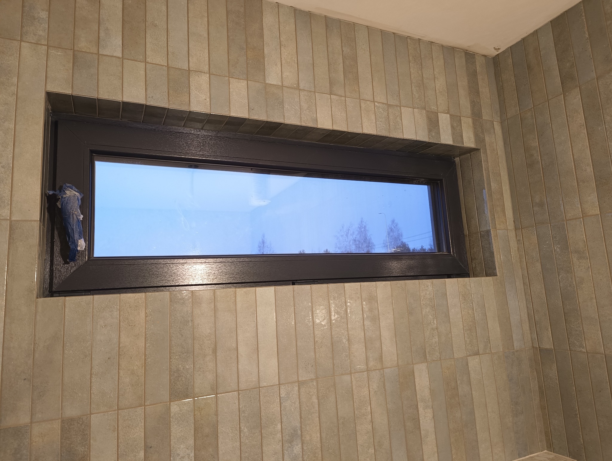 Wykończona ściana w łazience z beżowymi, wąskimi płytkami i oknem z ciemną ramą. Fragment folii ochronnej na ramie okna.