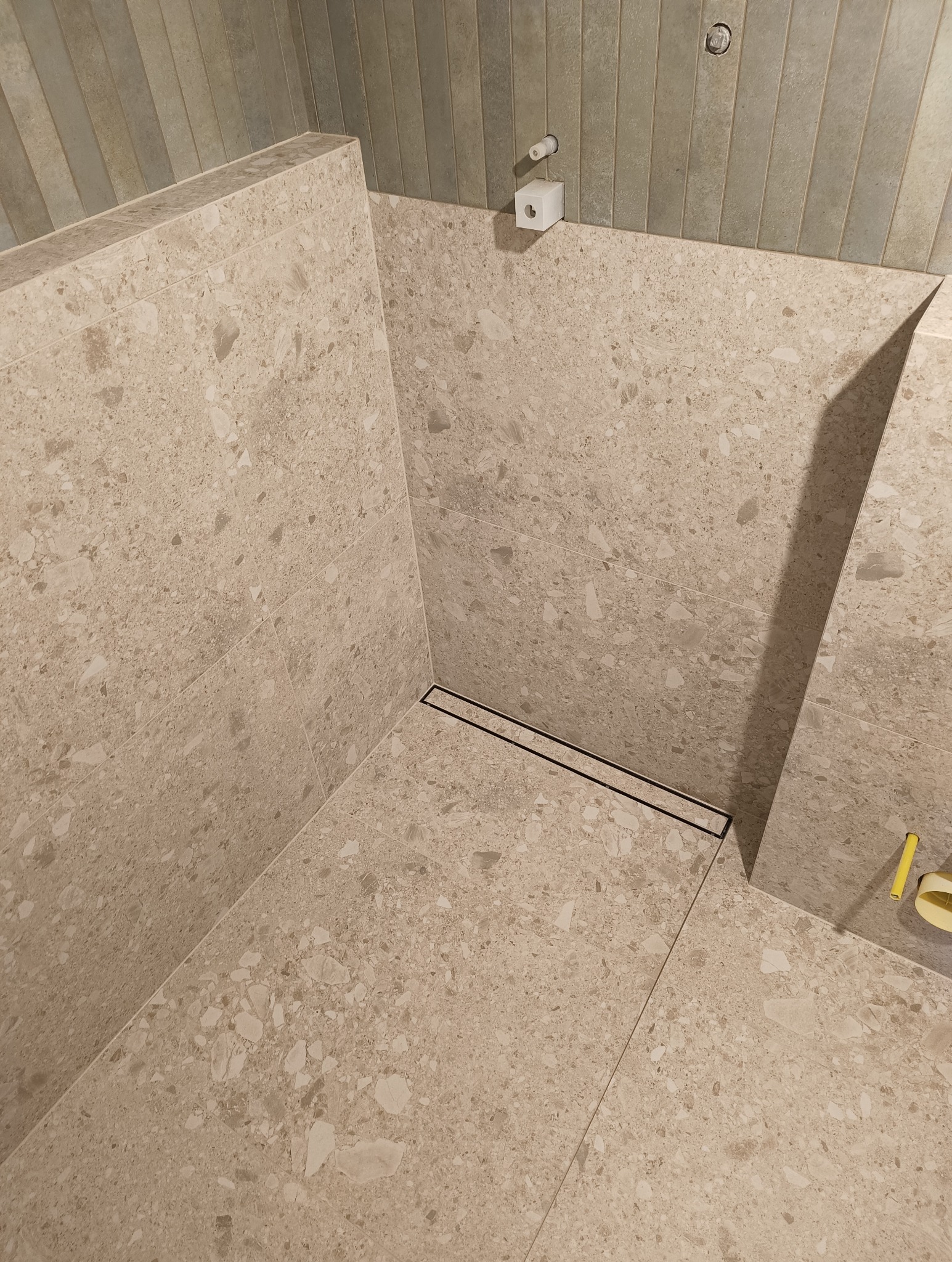 Beżowa kabina prysznicowa wyłożona płytkami imitującymi lastryko, z odpływem liniowym. Na ścianie widoczny dozownik mydła i uchwyt na papier toaletowy.