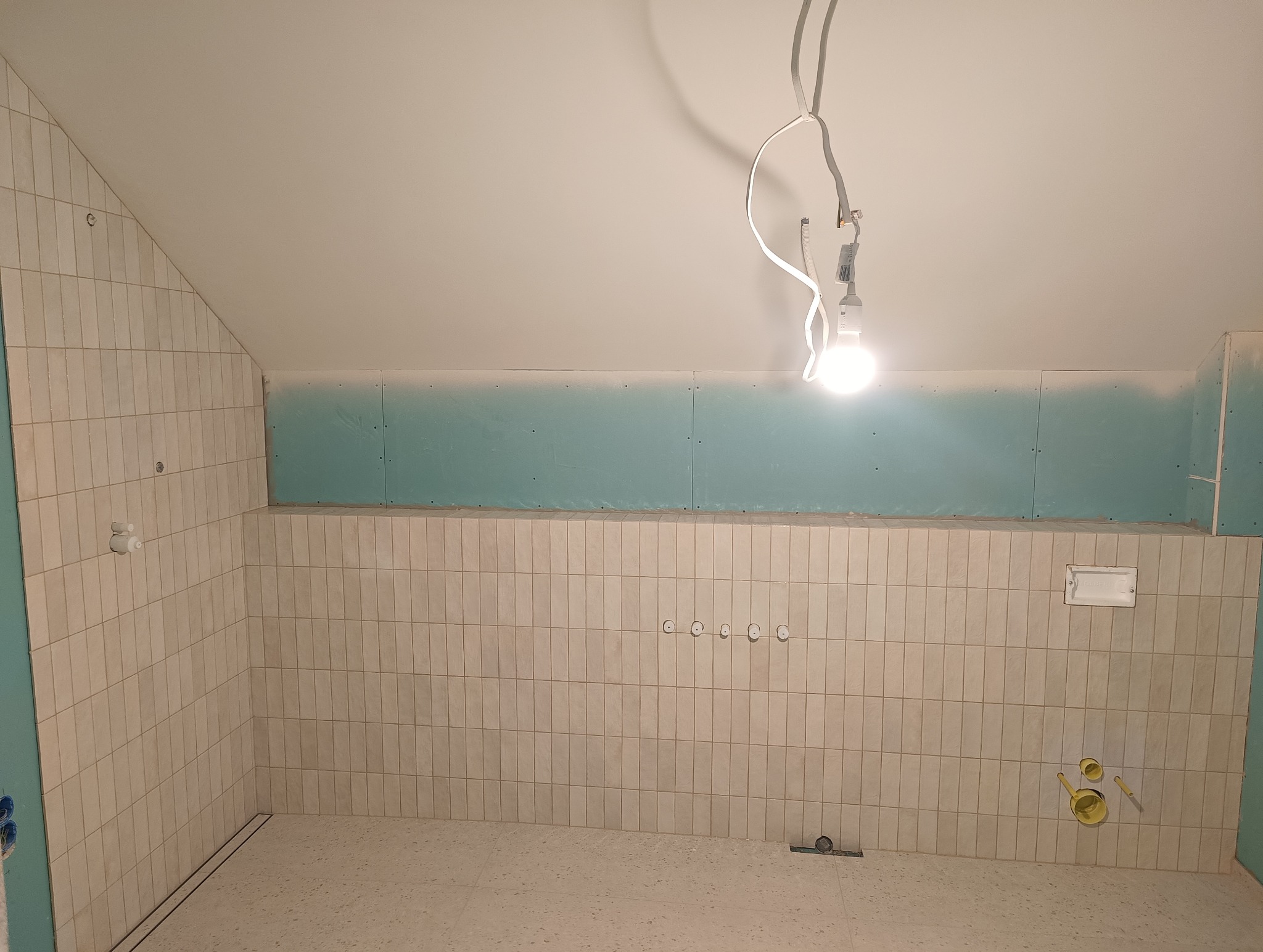 Łazienka na poddaszu w trakcie wykańczania: ułożone beżowe płytki na ścianach, płyty gipsowe na górze, widoczne instalacje i oświetlenie tymczasowe.