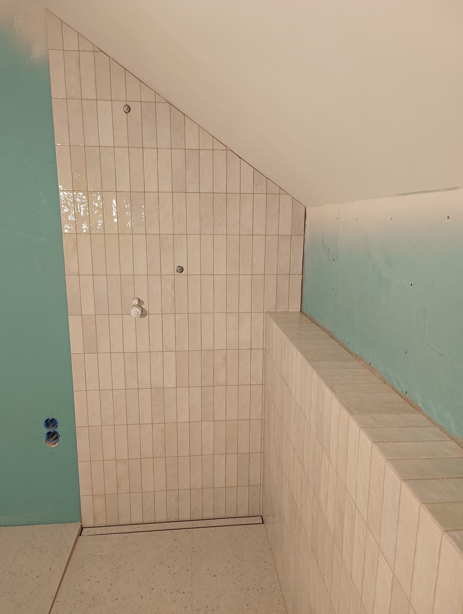 Wykończona beżowa glazura w kabinie prysznicowej ze ścianką, widoczny odpływ liniowy i fragment ściany z płyt gipsowych w kolorze zielonym.