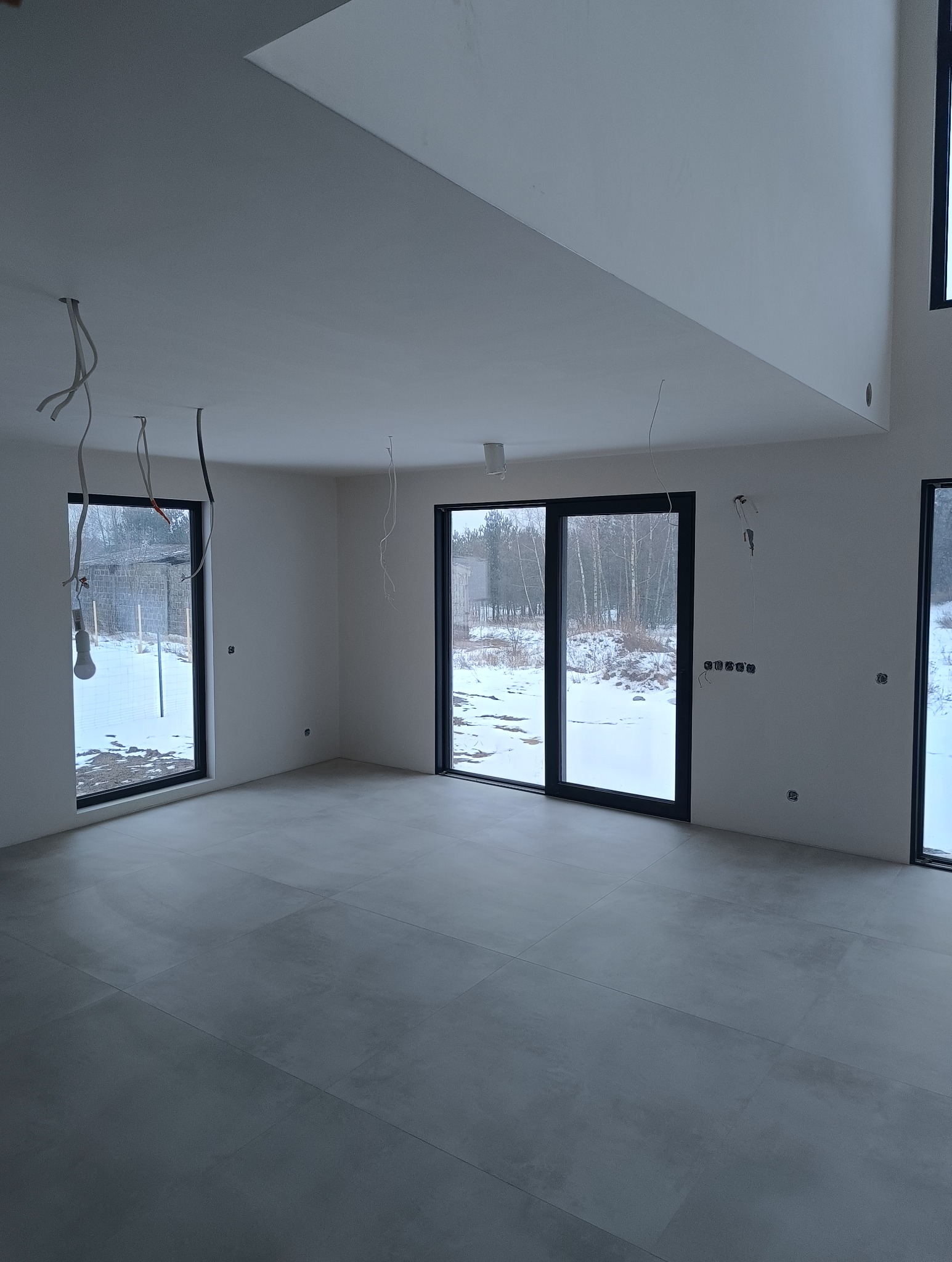 Szare płytki podłogowe w przestronnym, jasnym wnętrzu z dużymi oknami. Widoczne niedokończone instalacje elektryczne. Za oknami zimowy krajobraz.