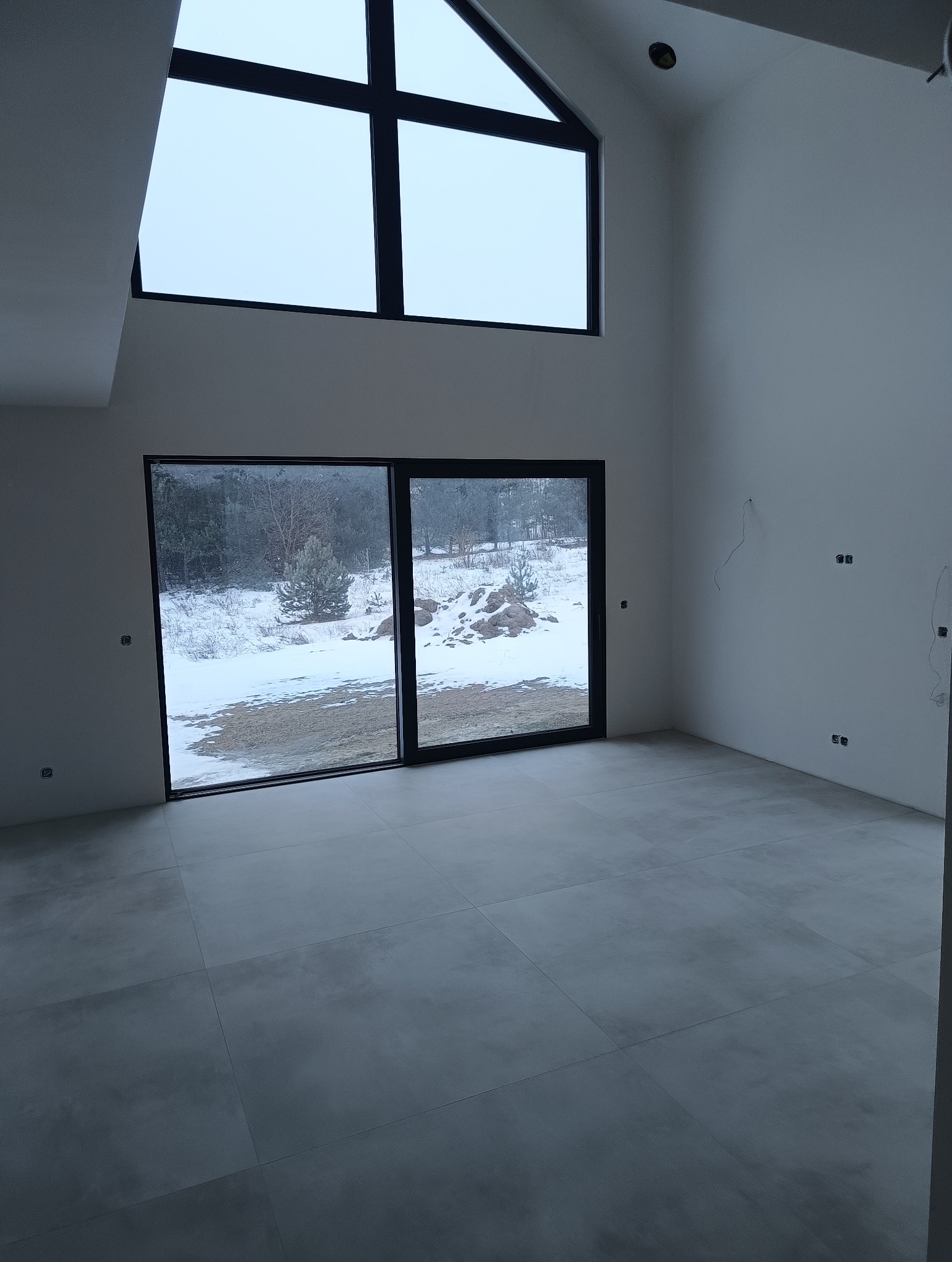 Minimalistyczne wnętrze z szarymi płytkami podłogowymi i dużymi oknami z widokiem na zimowy krajobraz. Surowe wykończenie ścian, widoczne punkty elektryczne.
