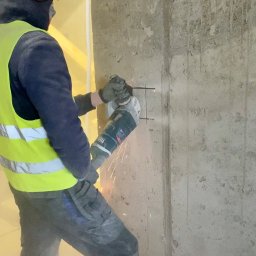 UPG Elektro - Elektryk w kamizelce tnie betonową ścianę szlifierką kątową, iskry lecą. Przygotowanie pod instalację elektryczną w surowym wnętrzu.