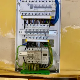 Rozdzielnica elektryczna w białej obudowie z zamontowanymi bezpiecznikami Eaton, przewodami i programatorem czasowym Orno, na tle ściany w trakcie remontu.