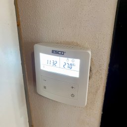 UPG Elektro - Biały termostat ESCO na teksturowanej ścianie, wskazujący 27 stopni Celsjusza. Widoczne przyciski sterowania i ikony trybów. Ujęcie z bliska, lekko z dołu.