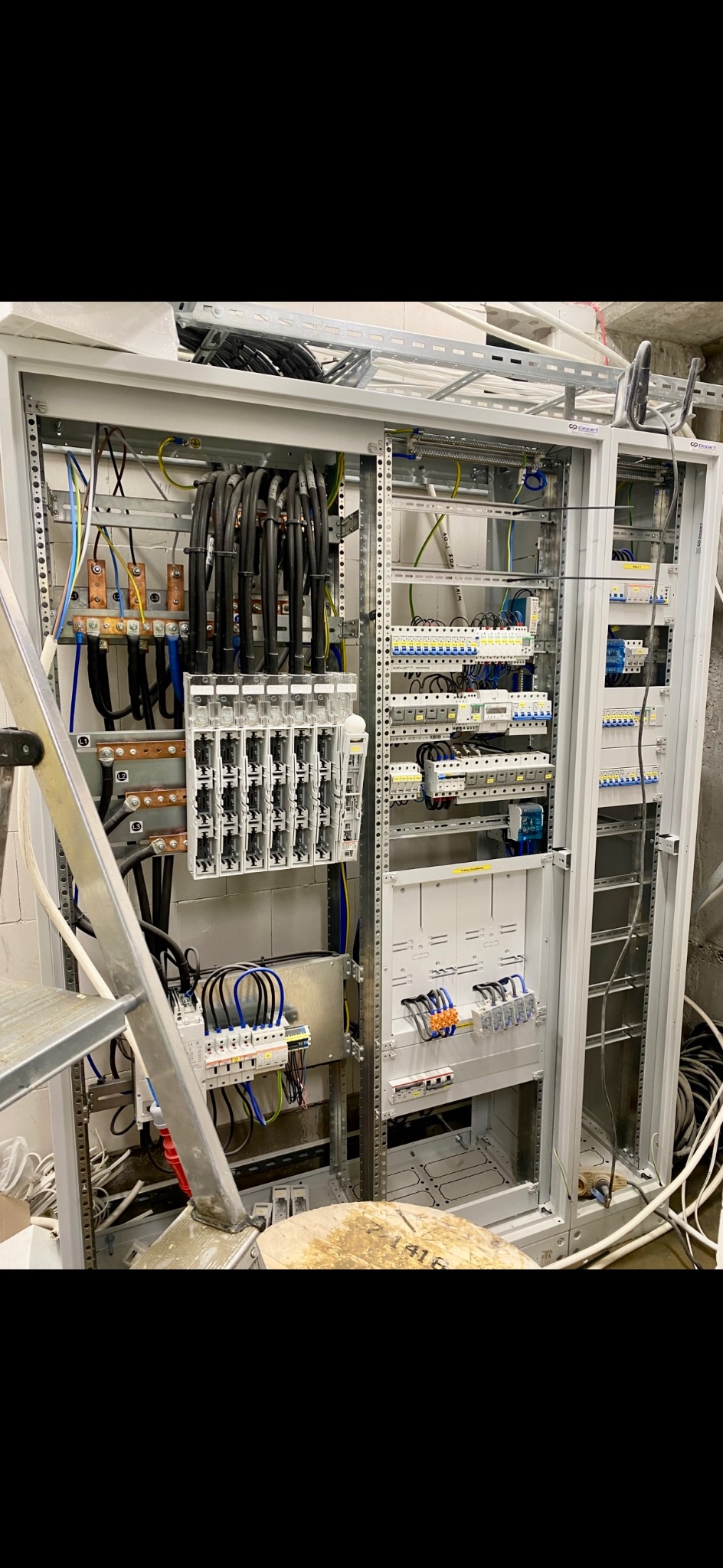 Rozdzielnia elektryczna w trakcie montażu: widoczne kable, bezpieczniki i elementy konstrukcyjne. Na pierwszym planie fragment drabiny.