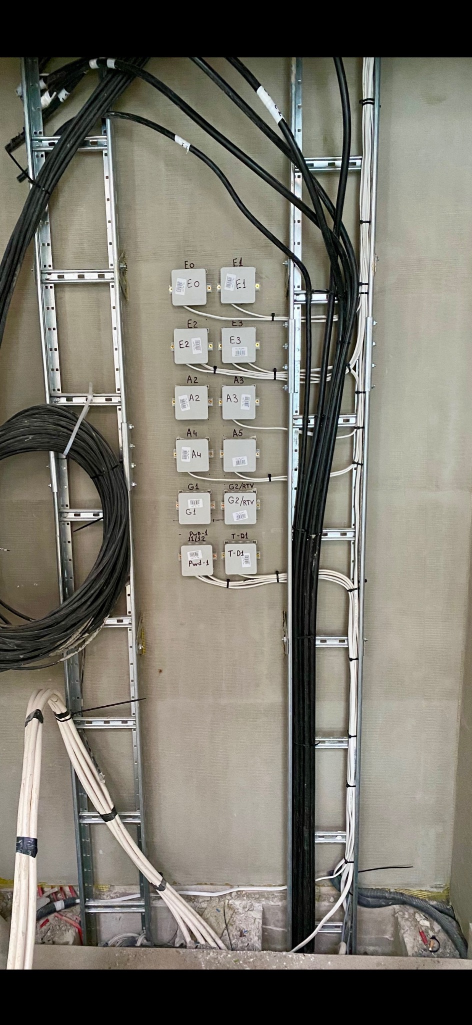 Szara ściana z zamontowanymi metalowymi profilami, w których poprowadzone są kable instalacji. Widoczne puszki z oznaczeniami E0-G2/RTV. Etap prac instalatorskich.