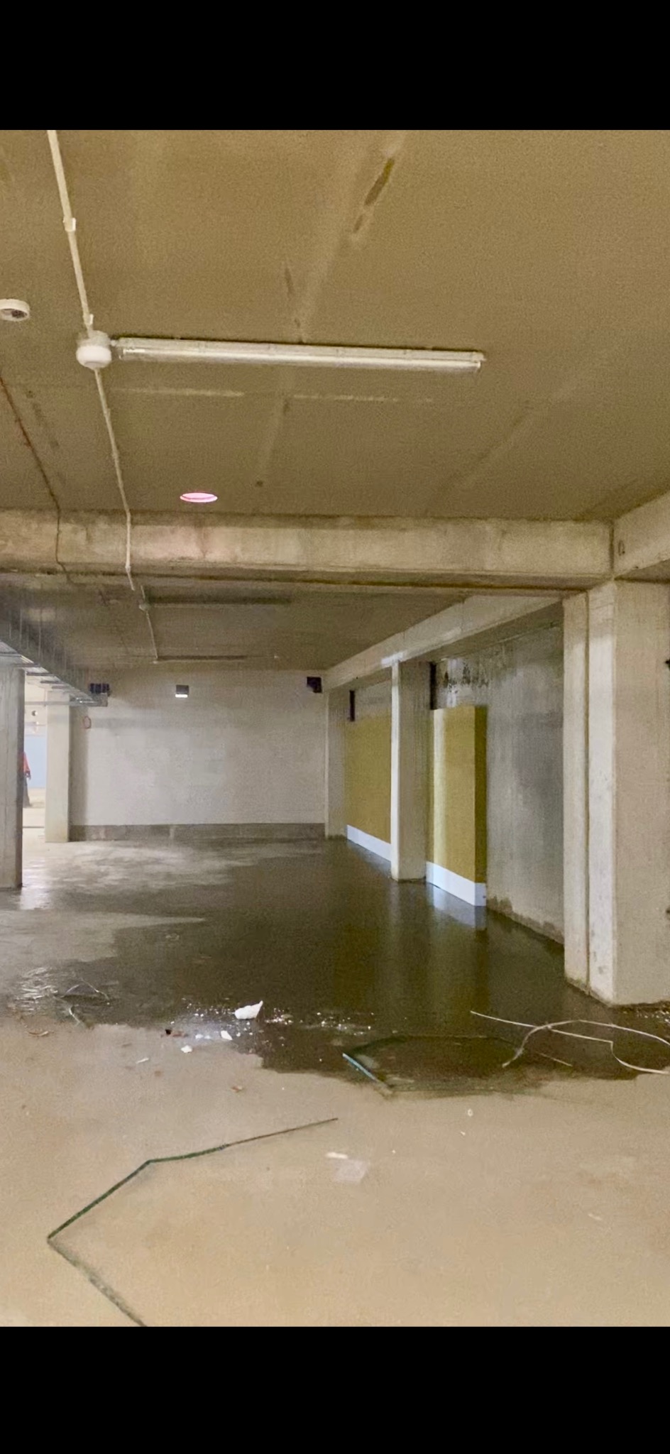 Wnętrze surowego pomieszczenia z widoczną instalacją elektryczną na suficie i elementami oświetlenia. Na podłodze woda i pozostałości po budowie.