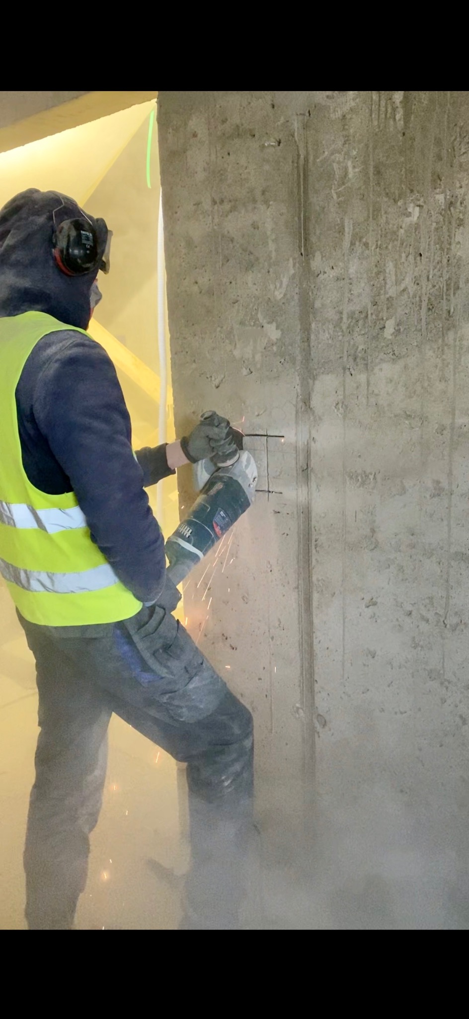 Elektryk w kamizelce tnie betonową ścianę szlifierką kątową, iskry lecą. Przygotowanie pod instalację elektryczną w surowym wnętrzu.