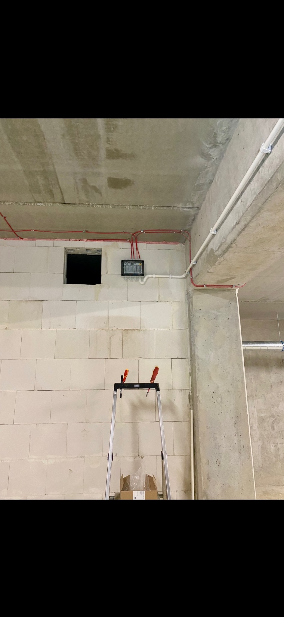 Rozdzielnica elektryczna z przewodami w czerwonej izolacji, zamontowana na ścianie z betonu komórkowego. Na pierwszym planie drabina z narzędziami.