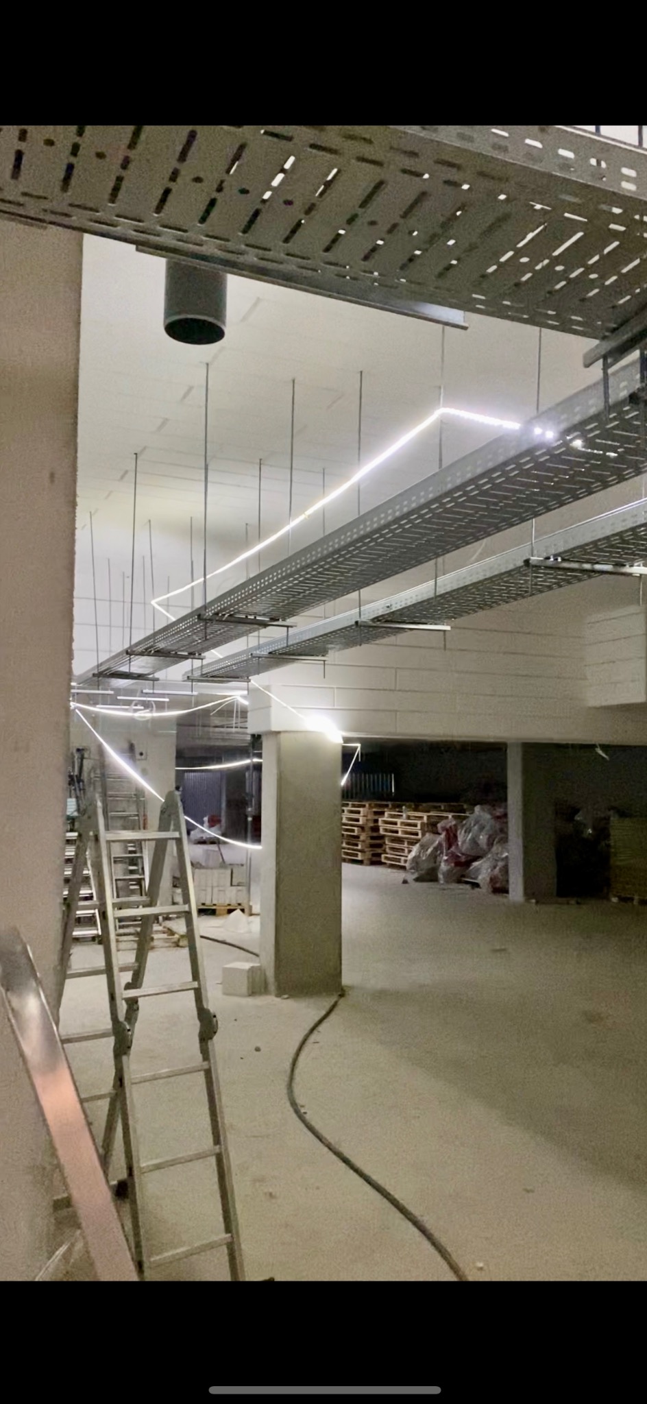 Nowoczesne oświetlenie LED zamontowane na metalowych korytkach kablowych pod sufitem w surowym wnętrzu. Widoczna drabina i elementy konstrukcyjne budynku.