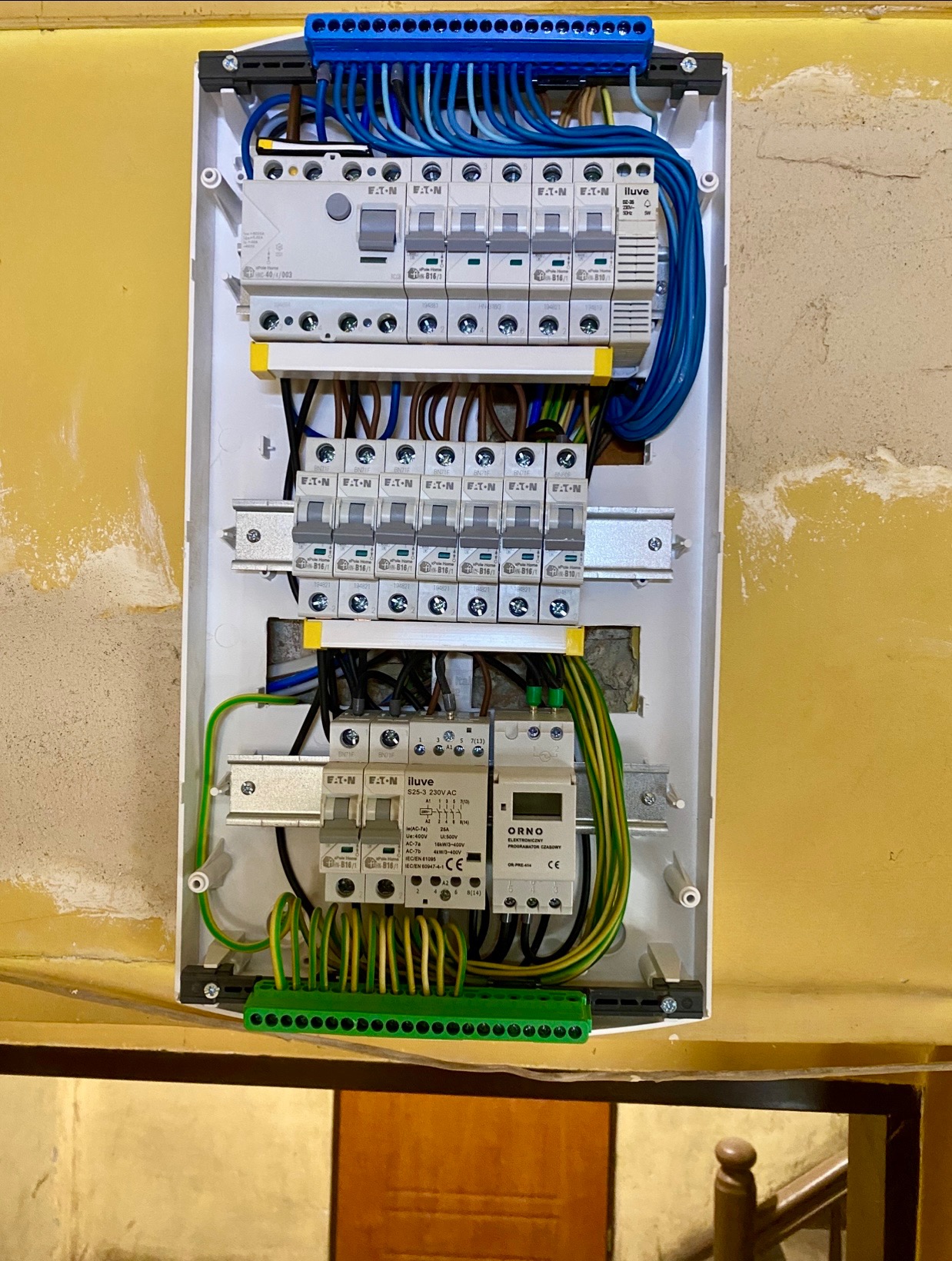 Rozdzielnica elektryczna w białej obudowie z zamontowanymi bezpiecznikami Eaton, przewodami i programatorem czasowym Orno, na tle ściany w trakcie remontu.