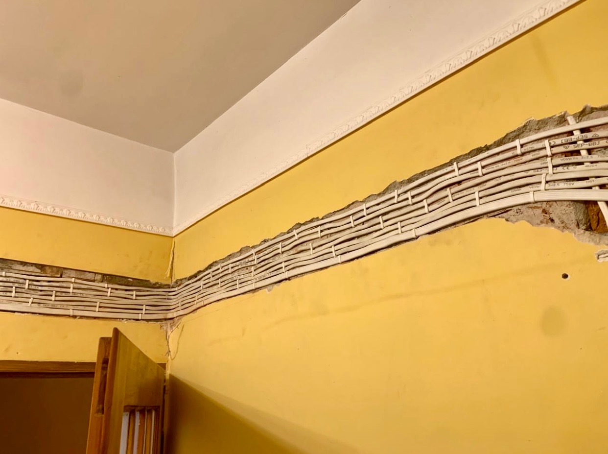 Żółta ściana z bruzdami, w których ułożono równolegle białe przewody elektryczne. Widoczne detale instalacji wewnątrz bruzd. Biały sufit z dekoracyjną listwą.