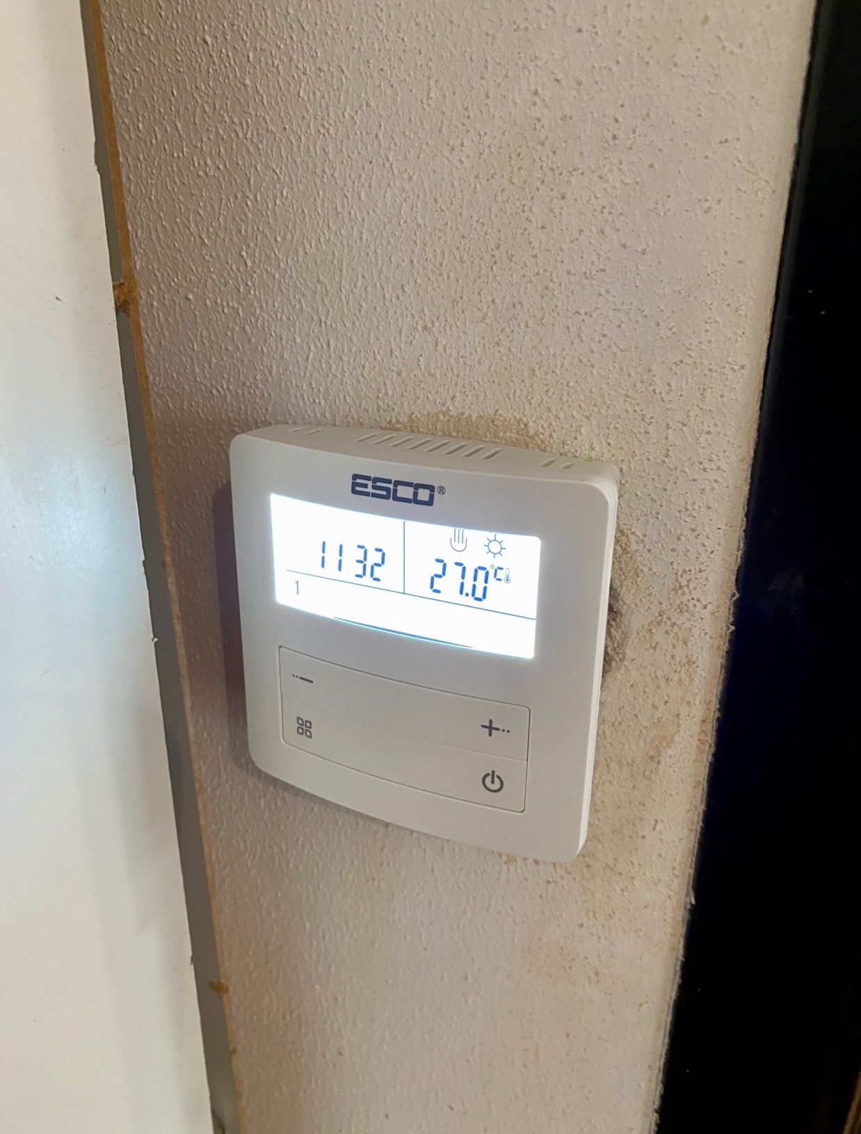Biały termostat ESCO na teksturowanej ścianie, wskazujący 27 stopni Celsjusza. Widoczne przyciski sterowania i ikony trybów. Ujęcie z bliska, lekko z dołu.