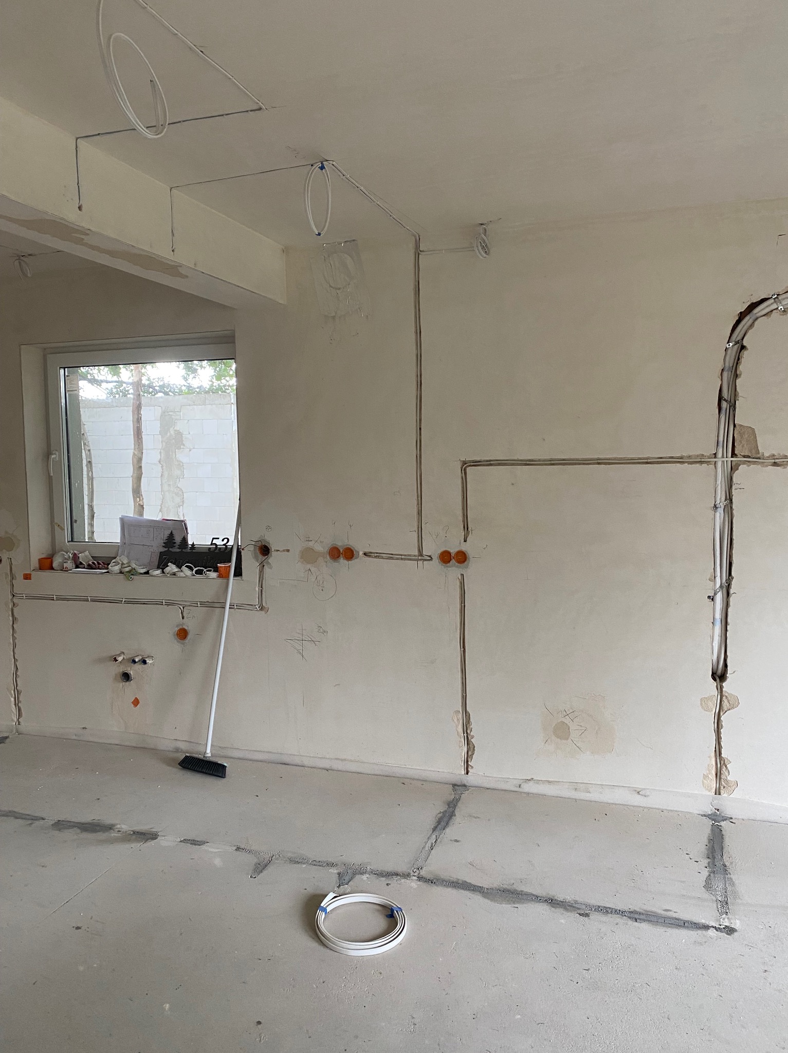 Wnętrze w trakcie prac elektrycznych: kable prowadzone w bruzdach ścian, puszki instalacyjne, zwój kabli na podłodze, widok na okno z planami i narzędziami.