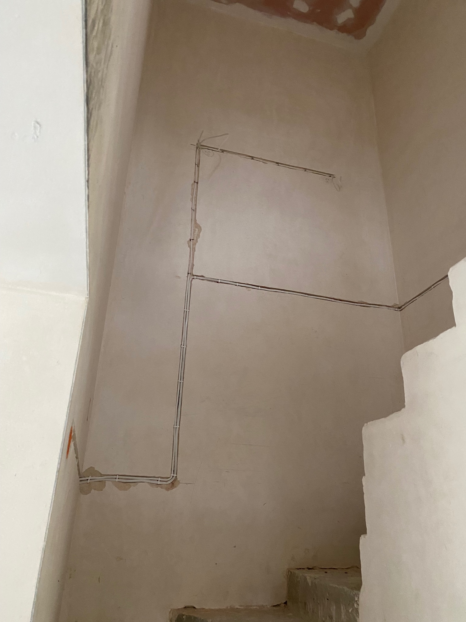 Kable elektryczne ułożone na ścianie w surowym wnętrzu w trakcie prac wykończeniowych. Widoczne białe rurki z przewodami, surowa ściana i fragment schodów.