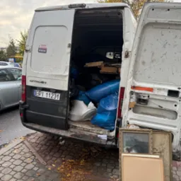 Otwarty, zapakowany bus marki Citroen z widocznymi workami, meblami i obrazami w ramach. Widok z tyłu na chodnik z liśćmi i fragment ulicy w tle.