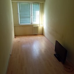 Grzegorz Mitura - Pusty pokój z parkietem i oknem z żaluzjami, przygotowany do opróżnienia. Na podłodze stoi telewizor. Ściany w odcieniach żółci, widoczne ślady użytkowania.