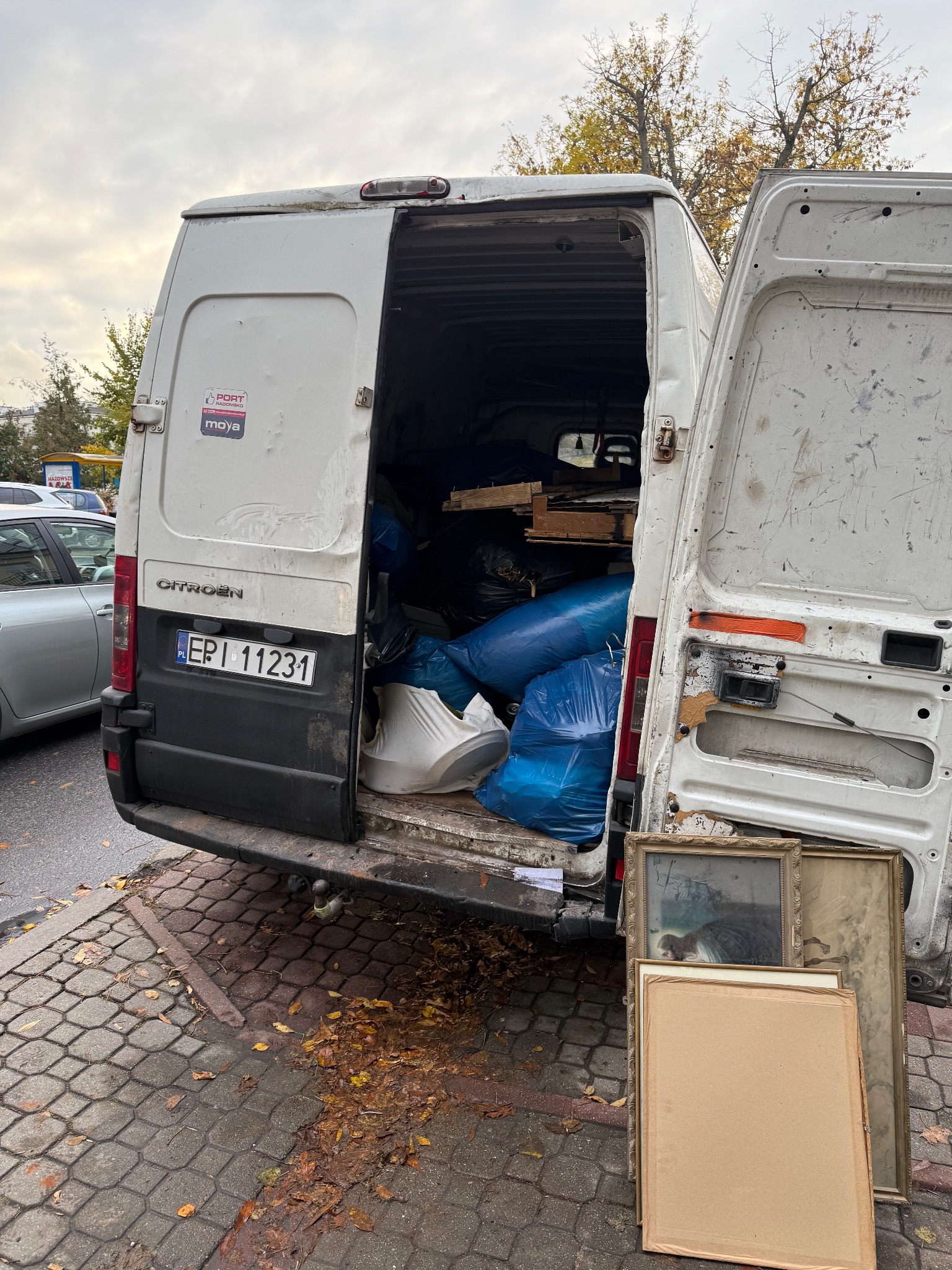 Otwarty, zapakowany bus marki Citroen z widocznymi workami, meblami i obrazami w ramach. Widok z tyłu na chodnik z liśćmi i fragment ulicy w tle.