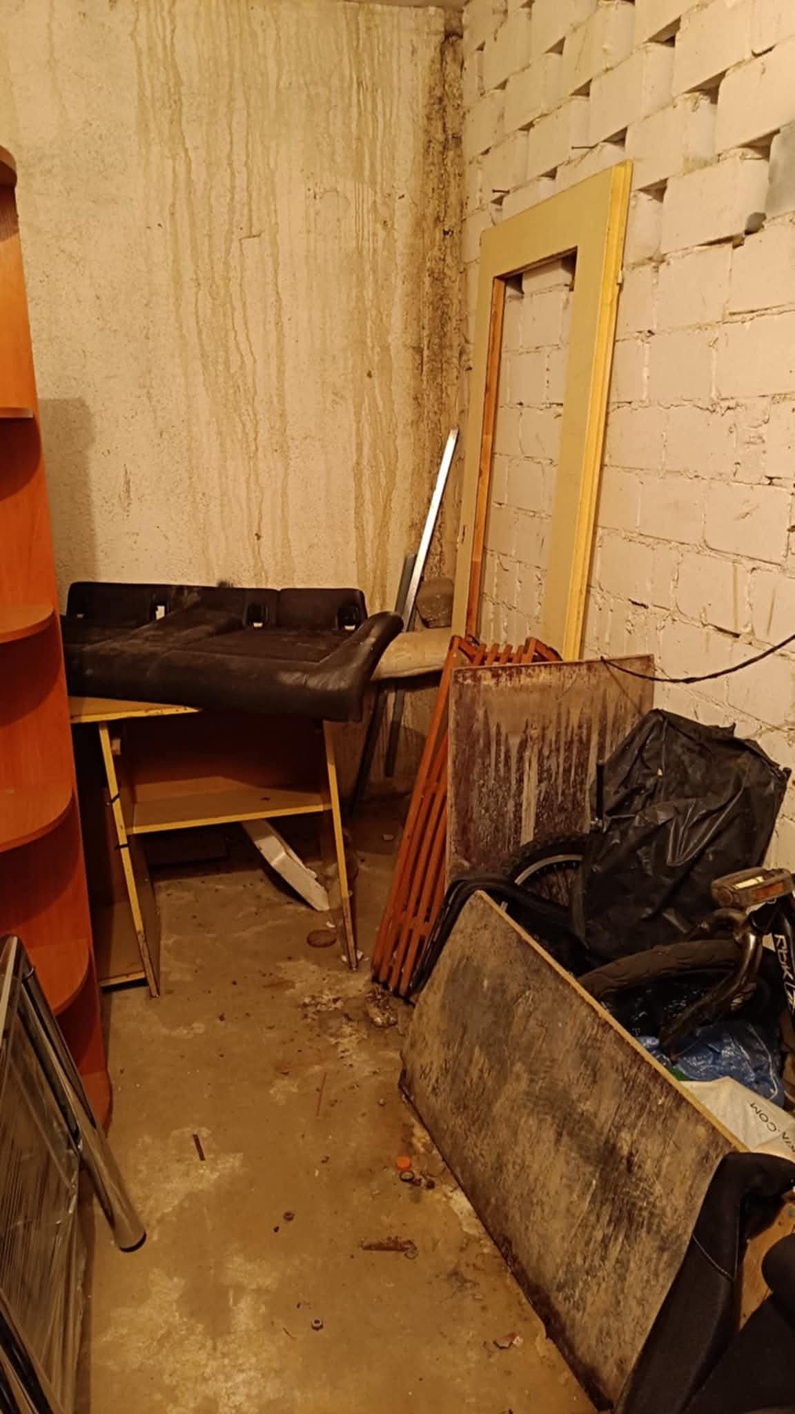 Zagracone pomieszczenie z meblami, ramą drzwiową i workiem. Ściany z zaciekami. Chaos i nieporządek w piwnicy lub magazynie.