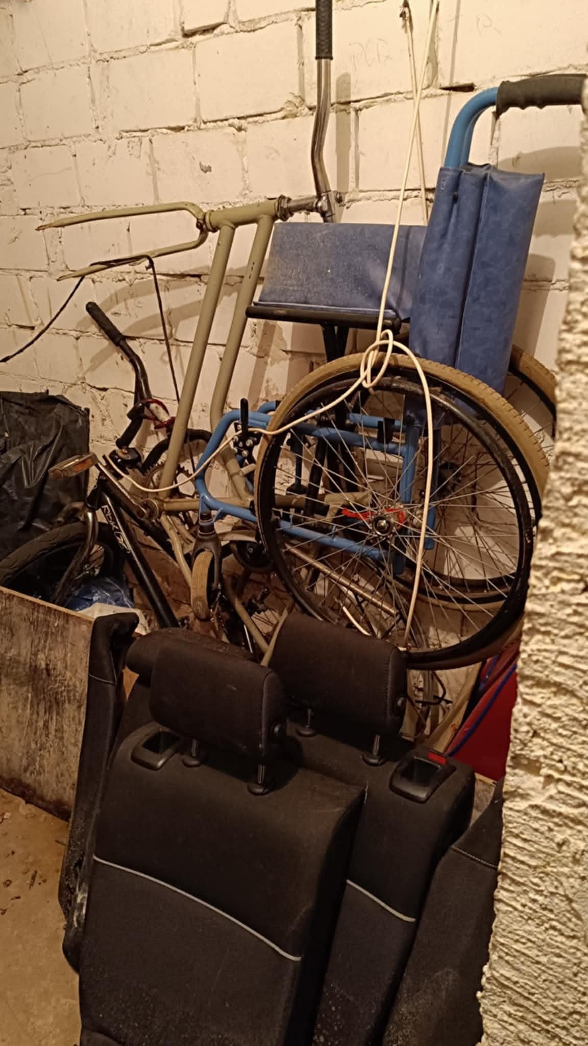 Stare rowery, wózek inwalidzki i fotele samochodowe stłoczone w rogu piwnicy z ceglaną ścianą. Chaos i zagracenie w pomieszczeniu.