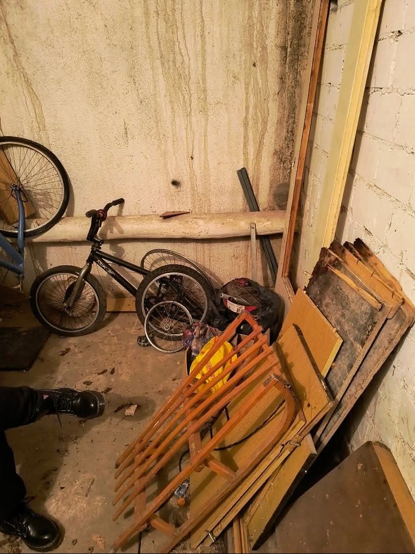 Zagracone pomieszczenie z rowerem, ramami okiennymi i innymi przedmiotami. Widoczne ślady wilgoci na ścianach. Niska jakość zdjęcia, słabe oświetlenie.